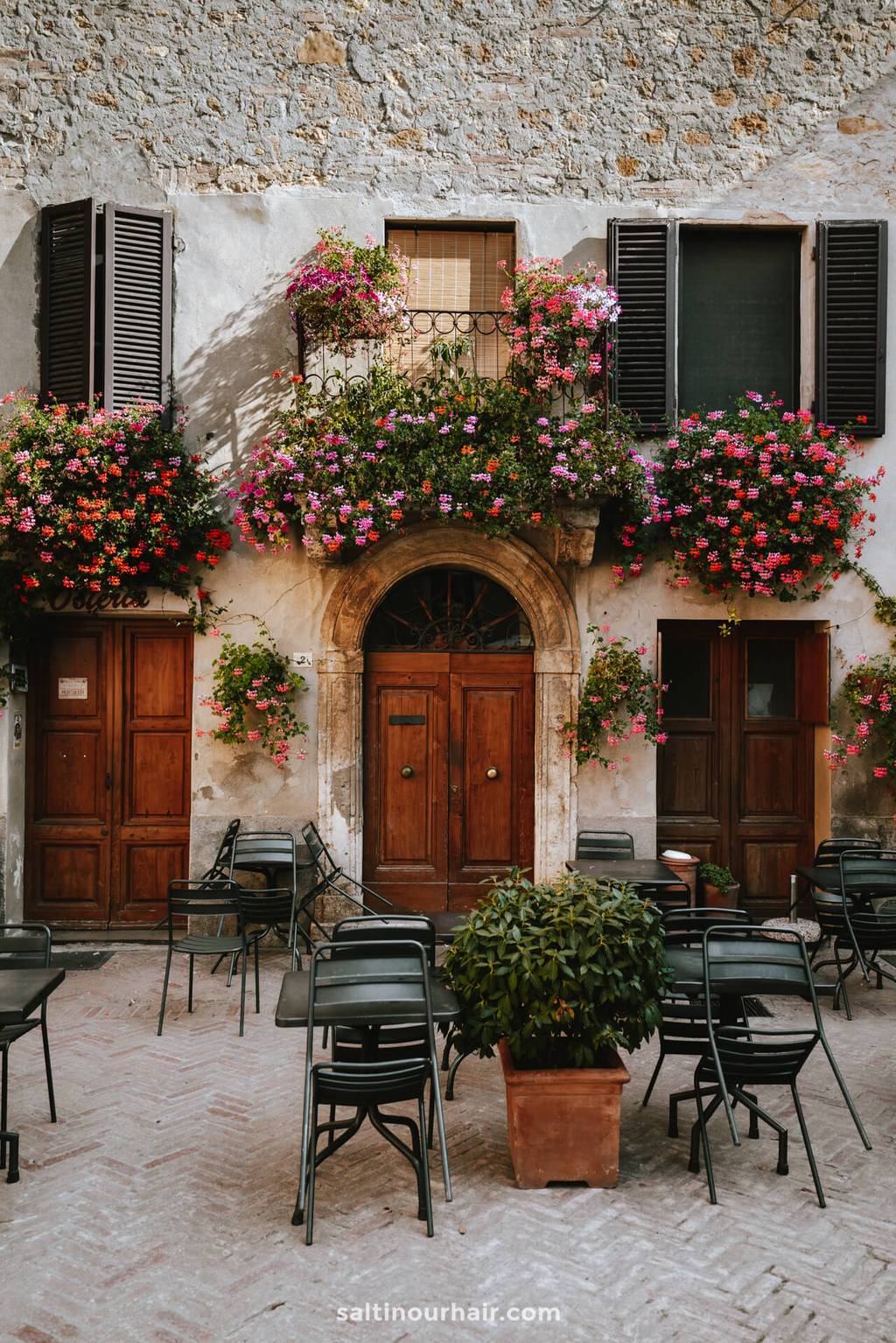A rustic stone building with wooden doors, shuttered windows, and balconies adorned with vibrant pink and red flowers—perfect for a pause during your Tuscany itinerary. Black chairs and tables are arranged outside.