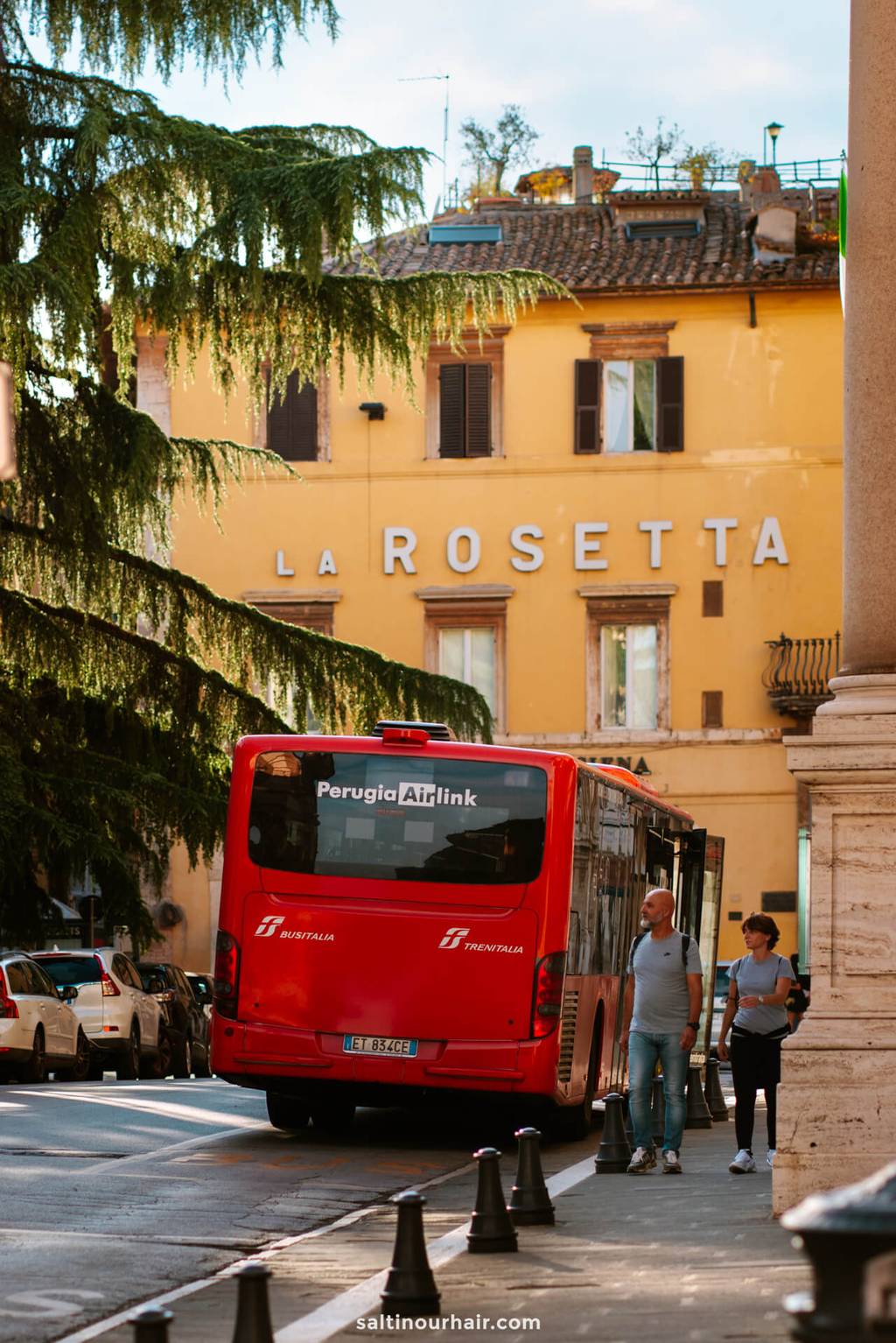 A red Perugia Airlink bus is parked on a street near people walking, with the La Rosetta hotel building in the background—a perfect scene to start your Tuscany itinerary.