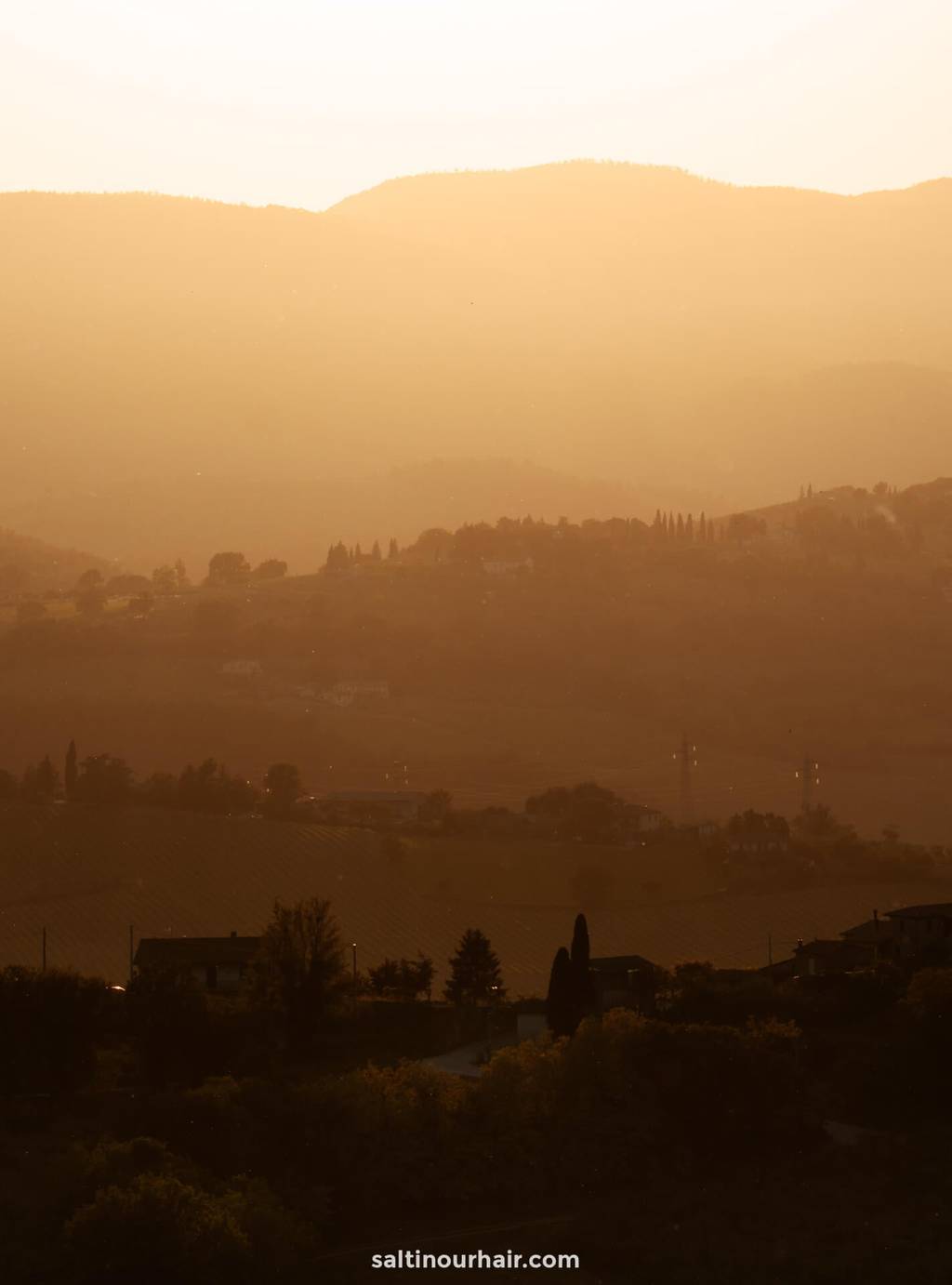 Hazy sunlight casts a golden glow over rolling hills and scattered trees, with faint outlines of houses—an ideal scene for any Tuscany itinerary.