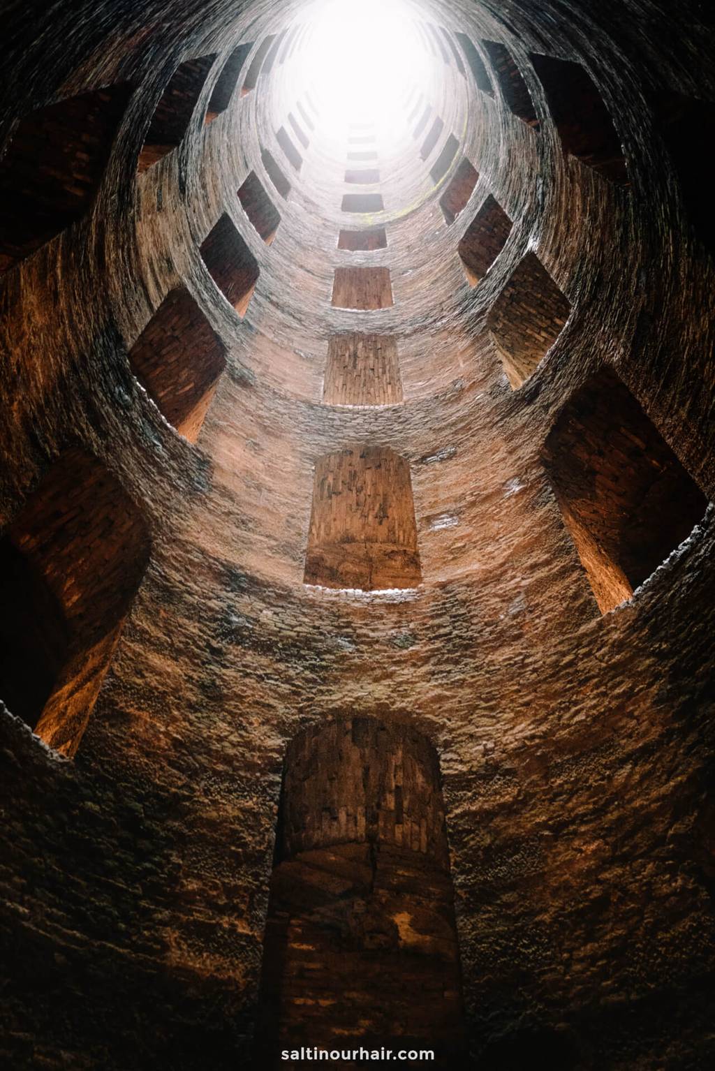 View looking up inside a cylindrical brick shaft with evenly spaced windows, leading to a bright light at the top—reminiscent of hidden gems on a Tuscany itinerary.