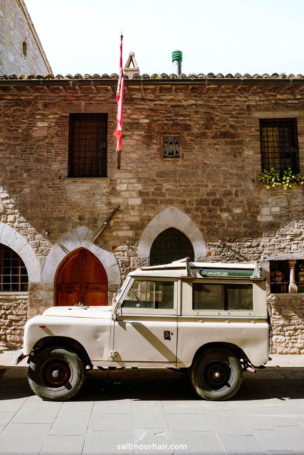 A vintage cream-colored Land Rover is parked on a sunny street in Rasiglia, Umbria, Italy, in front of an old stone building with arched doors and windows.