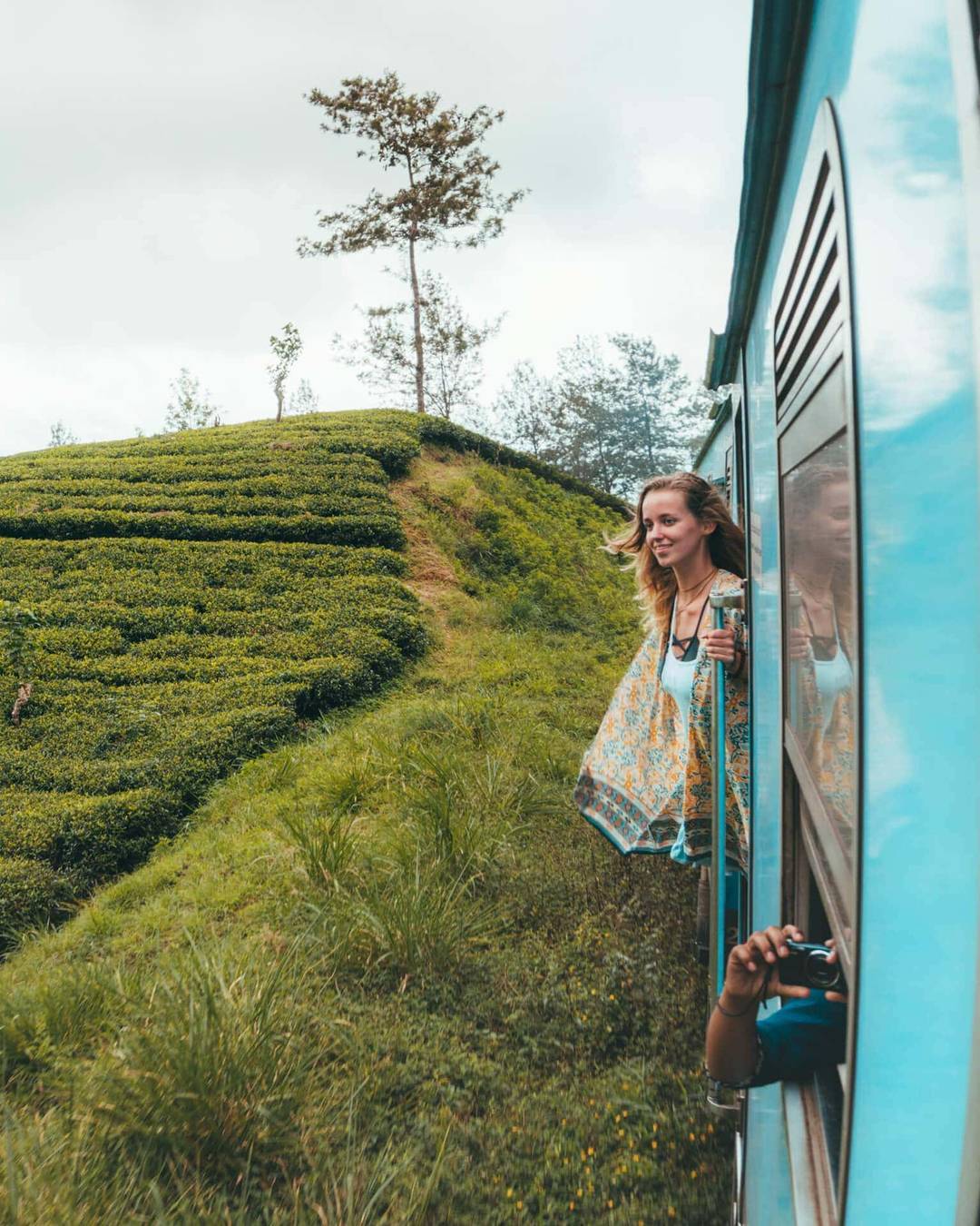 A woman leans out of a blue train window, smiling, as the iconic train from Kandy to Ella passes through green, hilly countryside with tea plantations in Sri Lanka.