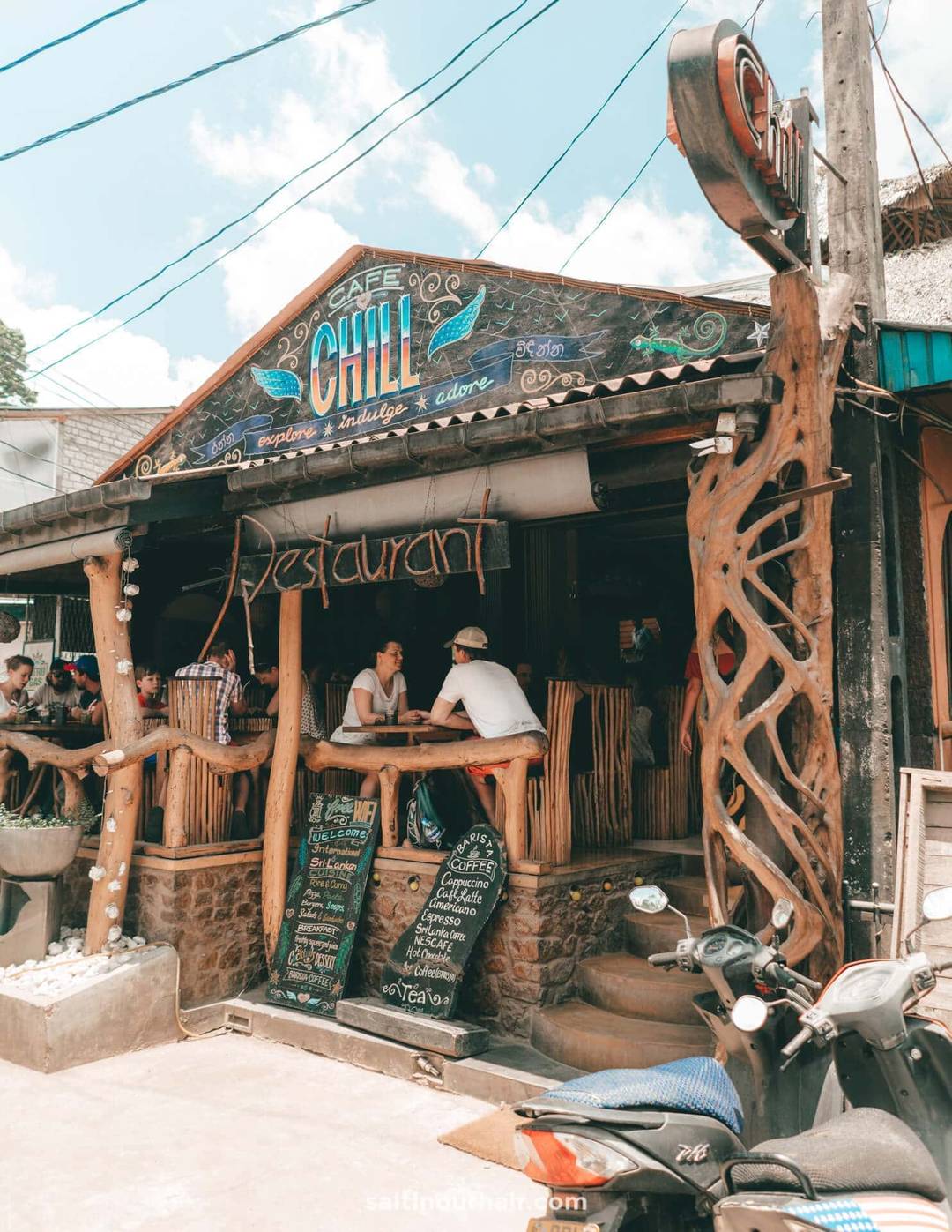 Small rustic café called Chill in Ella Town, Sri Lanka, with wooden decor, handwritten chalkboard menus, outdoor seating, and several people dining; scooters parked in front.