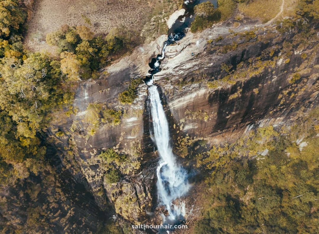 Aerial view of Diyaluma Falls, Sri Lanka, with its tall waterfall cascading down a steep rocky cliff surrounded by lush green and brown vegetation.