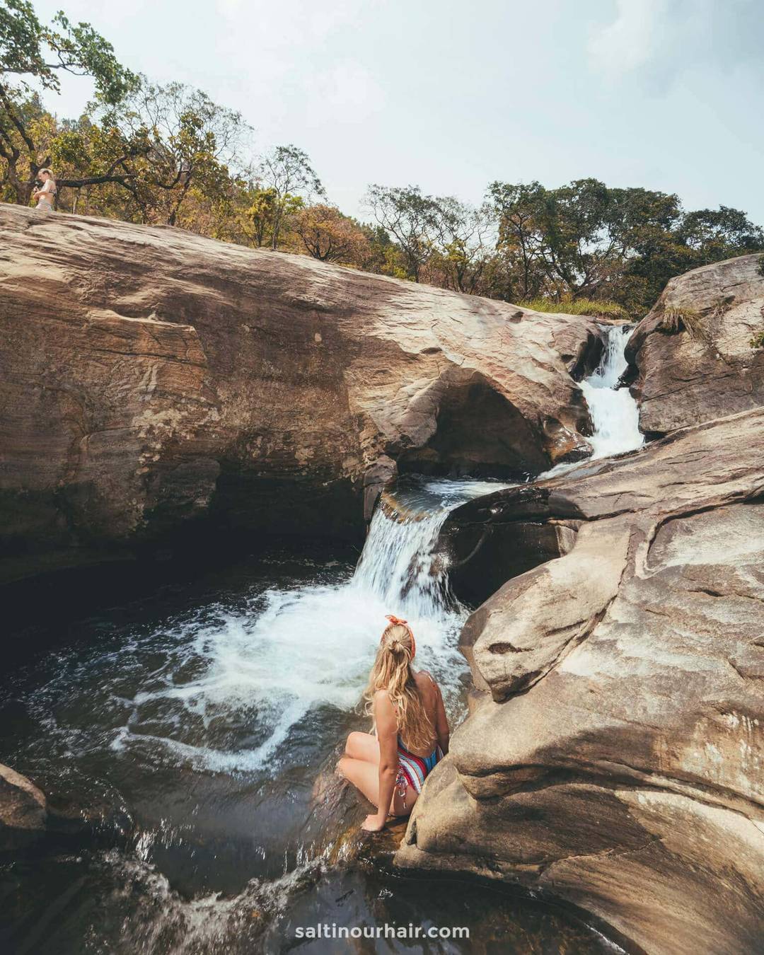 A woman sits on a rock by a small waterfall cascading into a pool, surrounded by large rock formations and trees&mdash;reminiscent of the serene scenery at Diyaluma Falls Sri Lanka.