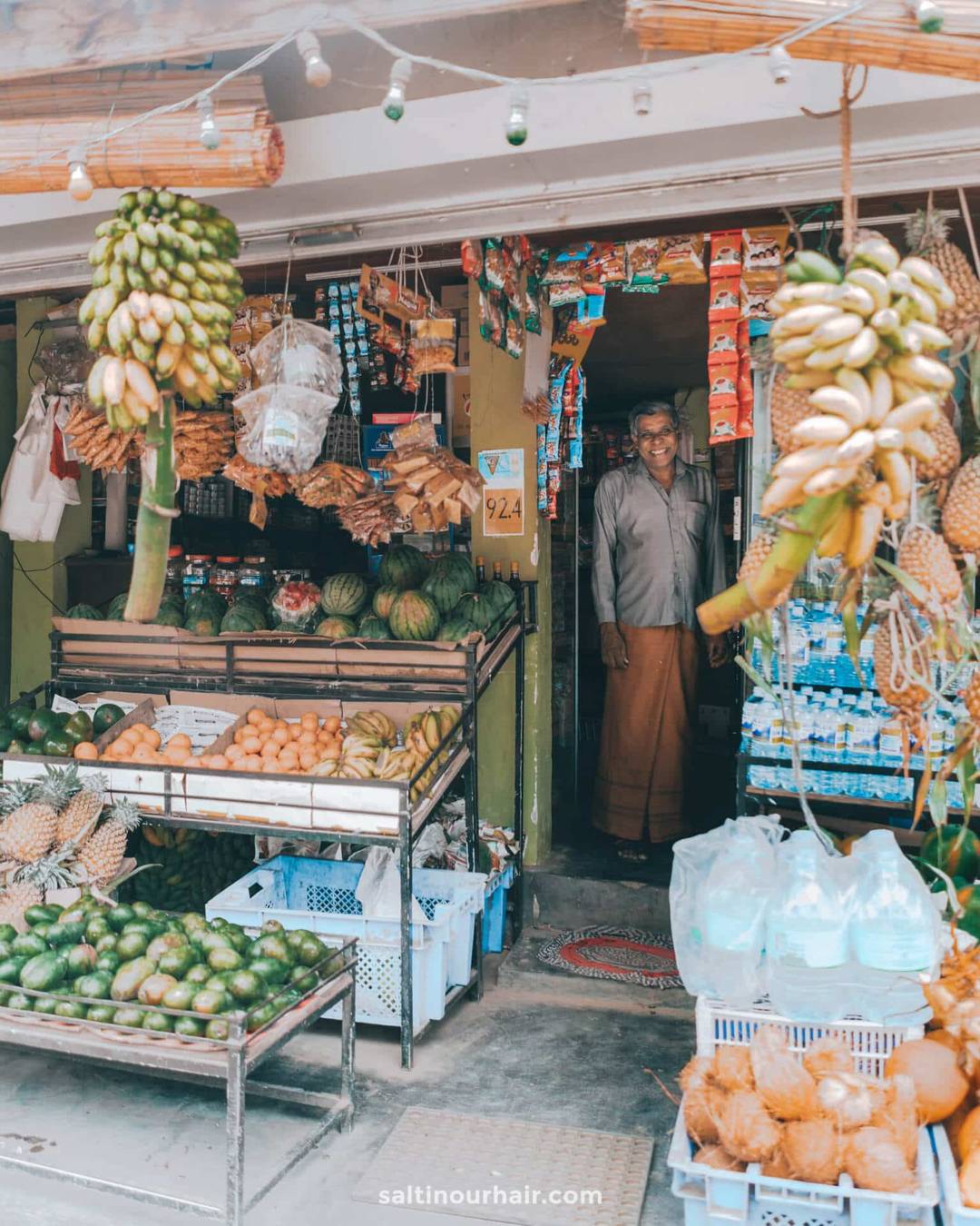 A shopkeeper stands in the entrance of a small produce store in Ella town, Sri Lanka, with bananas, watermelons, and other fruits and goods displayed outside.