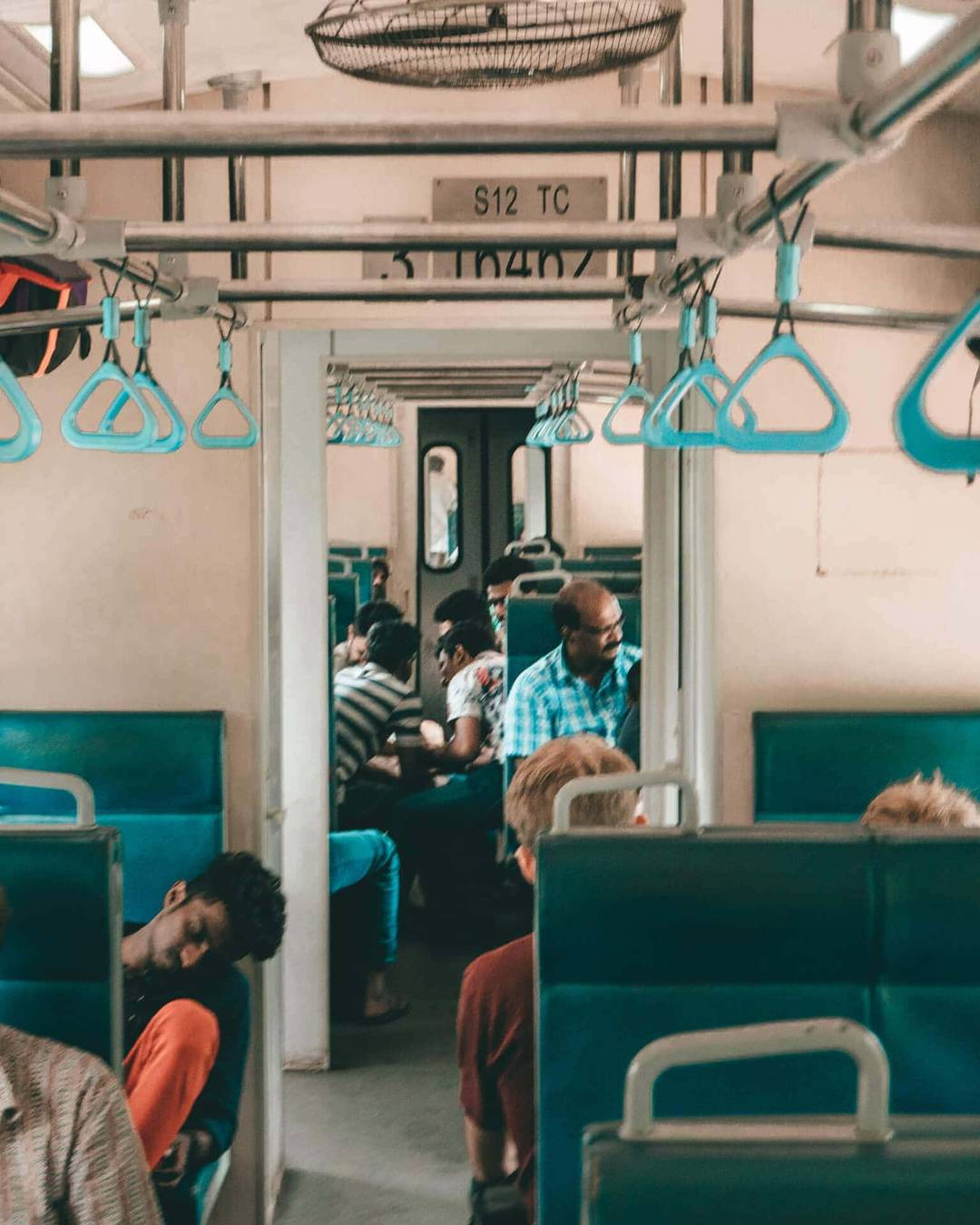 Passengers sit and rest on blue seats inside a train Sri Lanka carriage, with overhead handholds and open doors connecting to the next compartment.