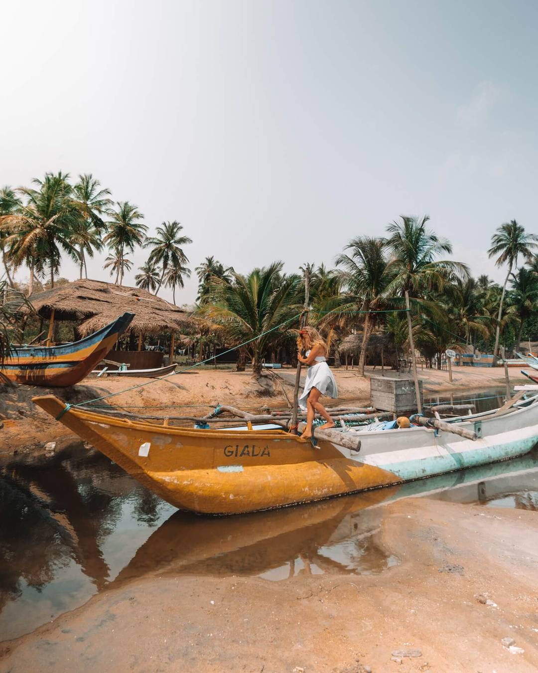 A person stands on a yellow fishing boat named GIALA near palm trees and huts on the sandy shore of Arugam Bay, Sri Lanka, with calm water in the background.
