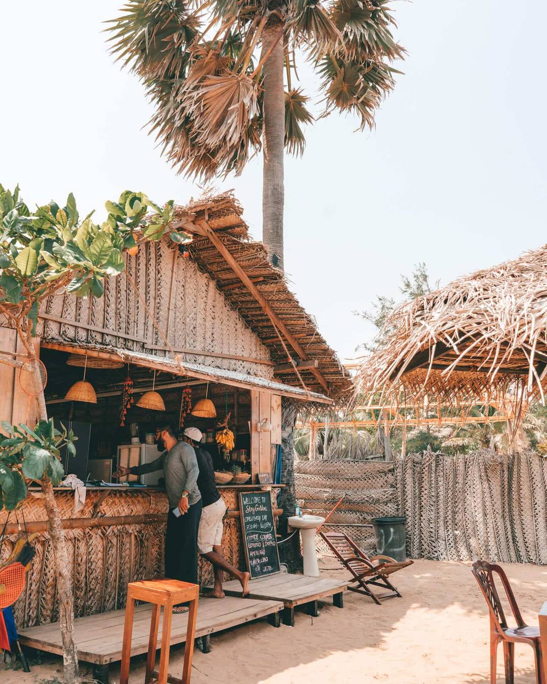 Two people stand at the counter of a thatched beachside caf&eacute; in Arugam Bay, Sri Lanka, surrounded by palm trees, with a chalkboard menu visible and outdoor seating on the sand.