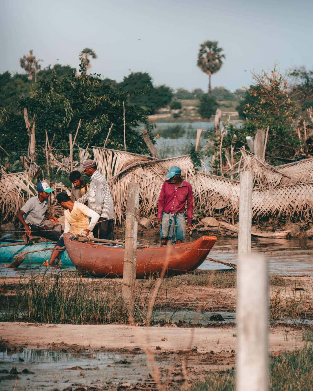 A group of people stand near small boats in a muddy, rural area surrounded by palm trees and wooden fences, capturing the authentic experience of a lagoon safari in Arugam Bay, Sri Lanka.