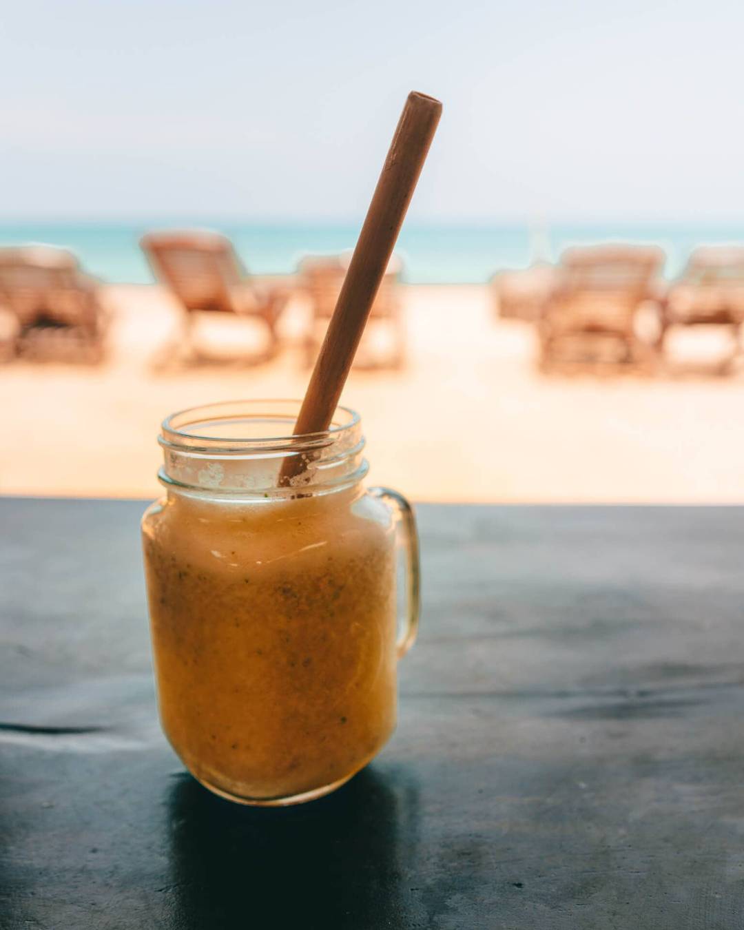 A mason jar filled with an orange smoothie and a straw sits on a table, with the sandy shores of Arugam Bay, Sri Lanka, and lounge chairs blurred in the background.