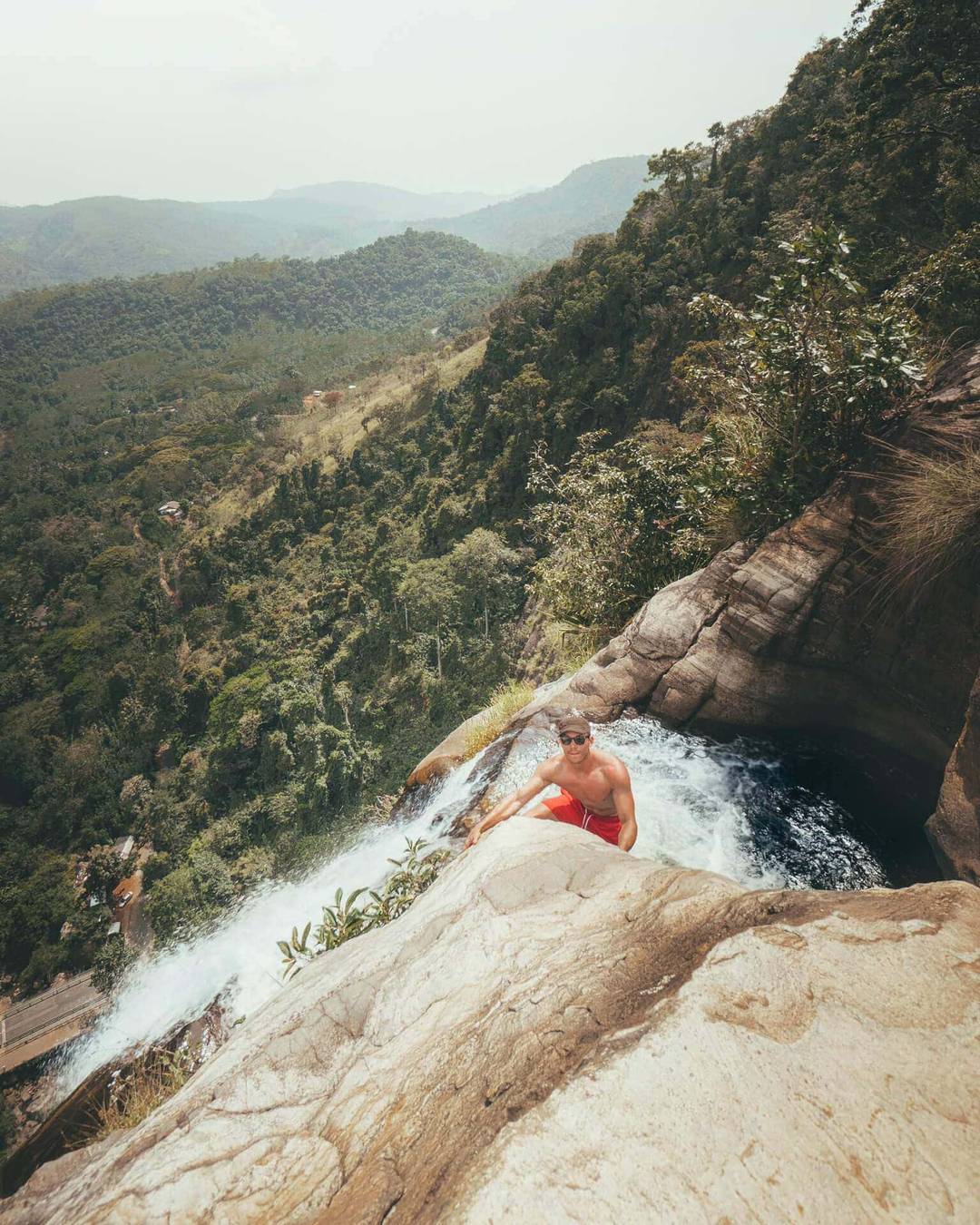 A person in a red swimsuit sits at the edge of Diyaluma Falls, Sri Lanka, on a rocky cliff, overlooking a lush green valley with distant hills.