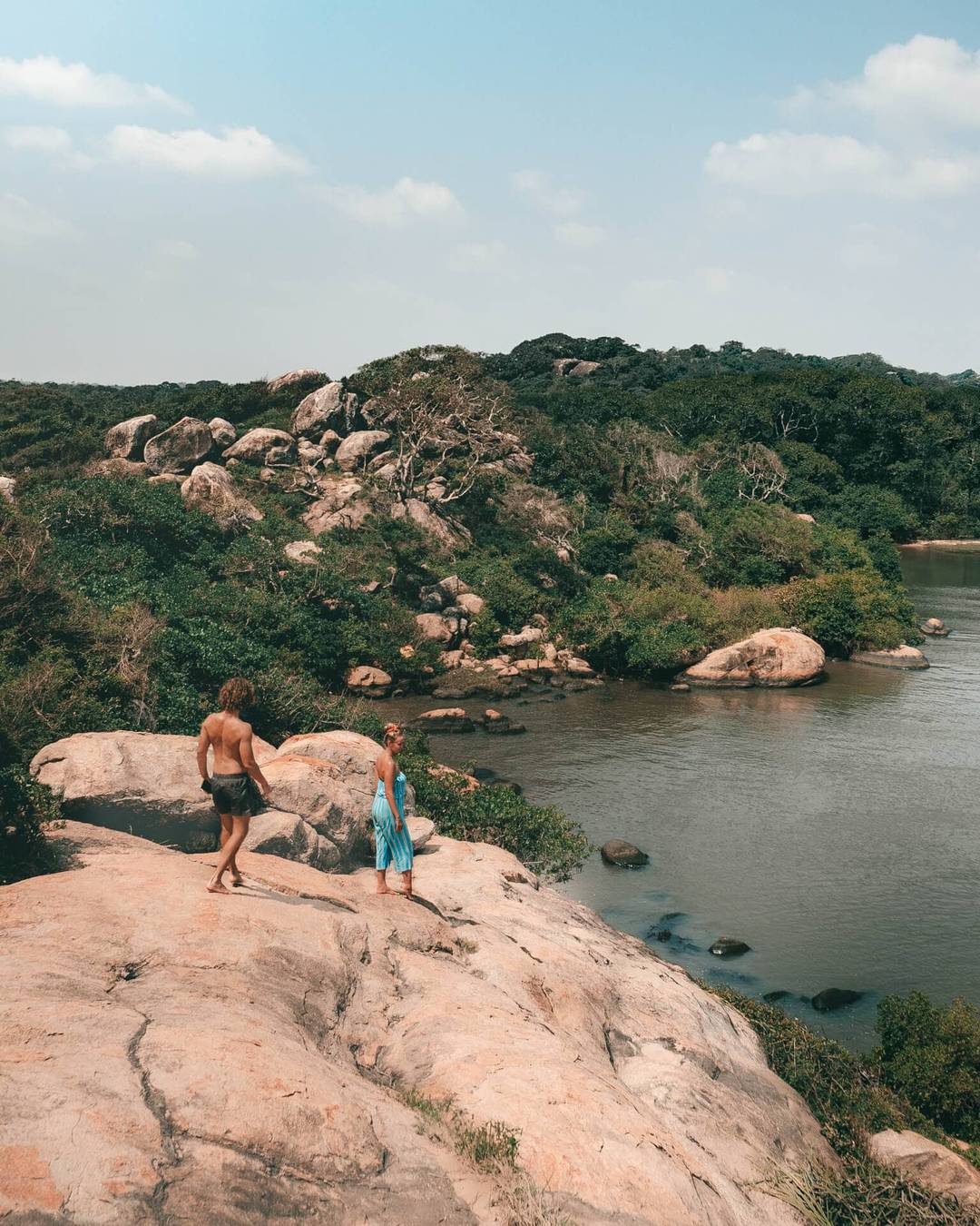 Two people stand on a rocky outcrop near a river, surrounded by green trees and hills under a partly cloudy sky, reminiscent of the scenic views at Elephant Rock in Arugam Bay, Sri Lanka.