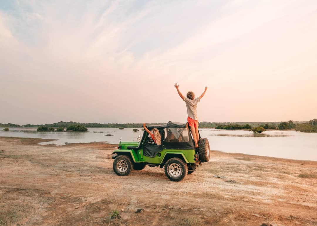 Two people are on and in a green off-road vehicle parked by a lake near Trincomalee, Sri Lanka; one stands on the back with arms raised, the other sits inside, surrounded by open water and distant greenery.