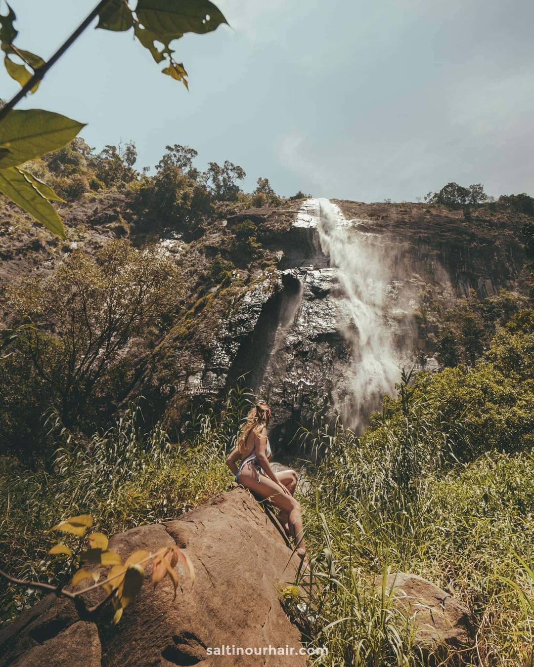 A woman in swimwear sits on a rock before Diyaluma Falls, Sri Lanka, surrounded by lush greenery and a rocky cliff beneath a partly cloudy sky.