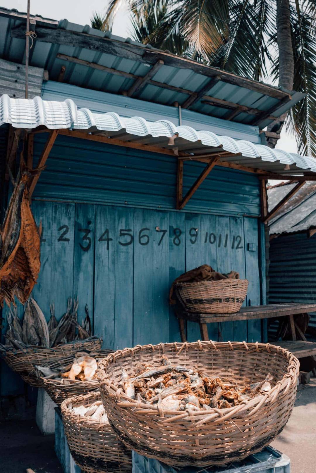 A blue wooden stall in Trincomalee, Sri Lanka, with numbers 1 to 12 painted on it, baskets of dried fish, and palm trees in the background.