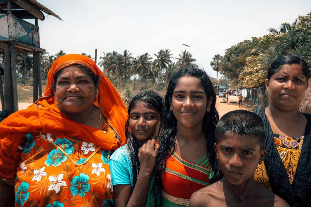 Five people, including three girls, a woman in an orange floral dress and headscarf, and a boy, stand outdoors with palm trees in the background on a sunny day in Trincomalee, Sri Lanka.