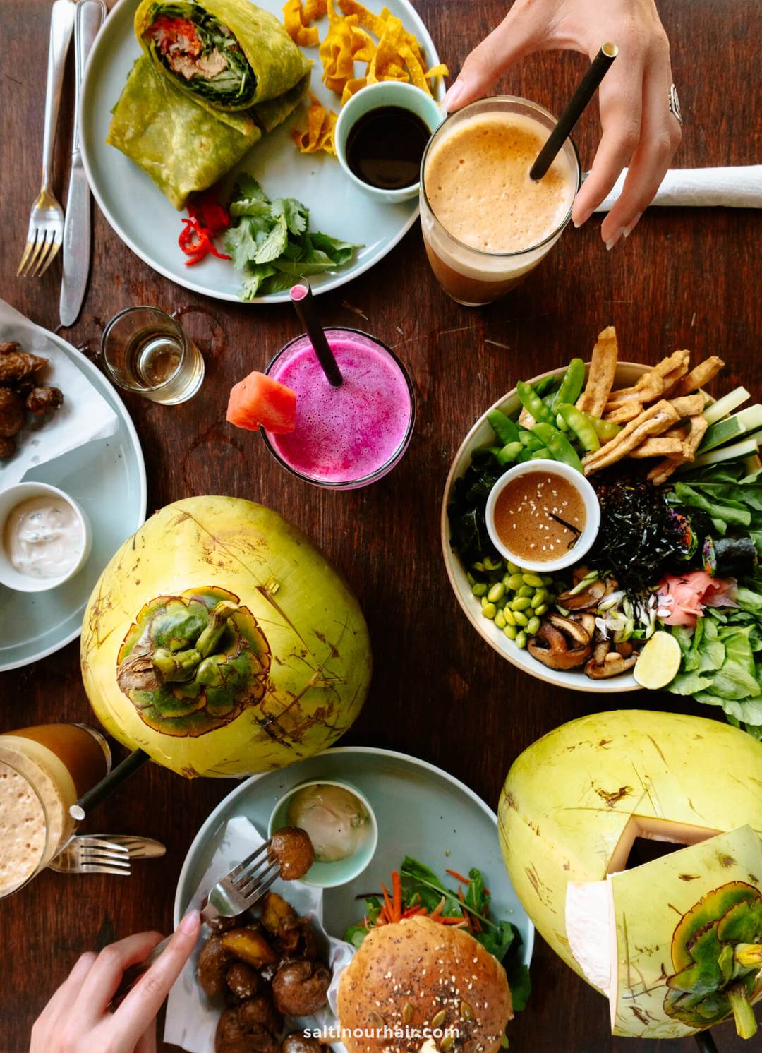 Top-down view of a table in Canggu Bali with fresh coconuts, vegetarian burgers, salads, wraps, smoothies, and two people reaching for drinks.