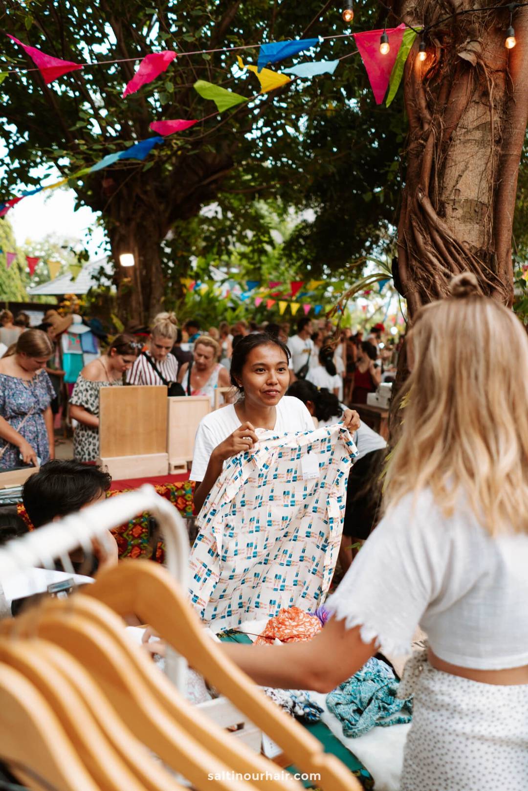 At La Brisa Sunday Market in Canggu, Bali, a woman holds up a patterned shirt while speaking to a vendor, with people browsing and colorful bunting overhead.