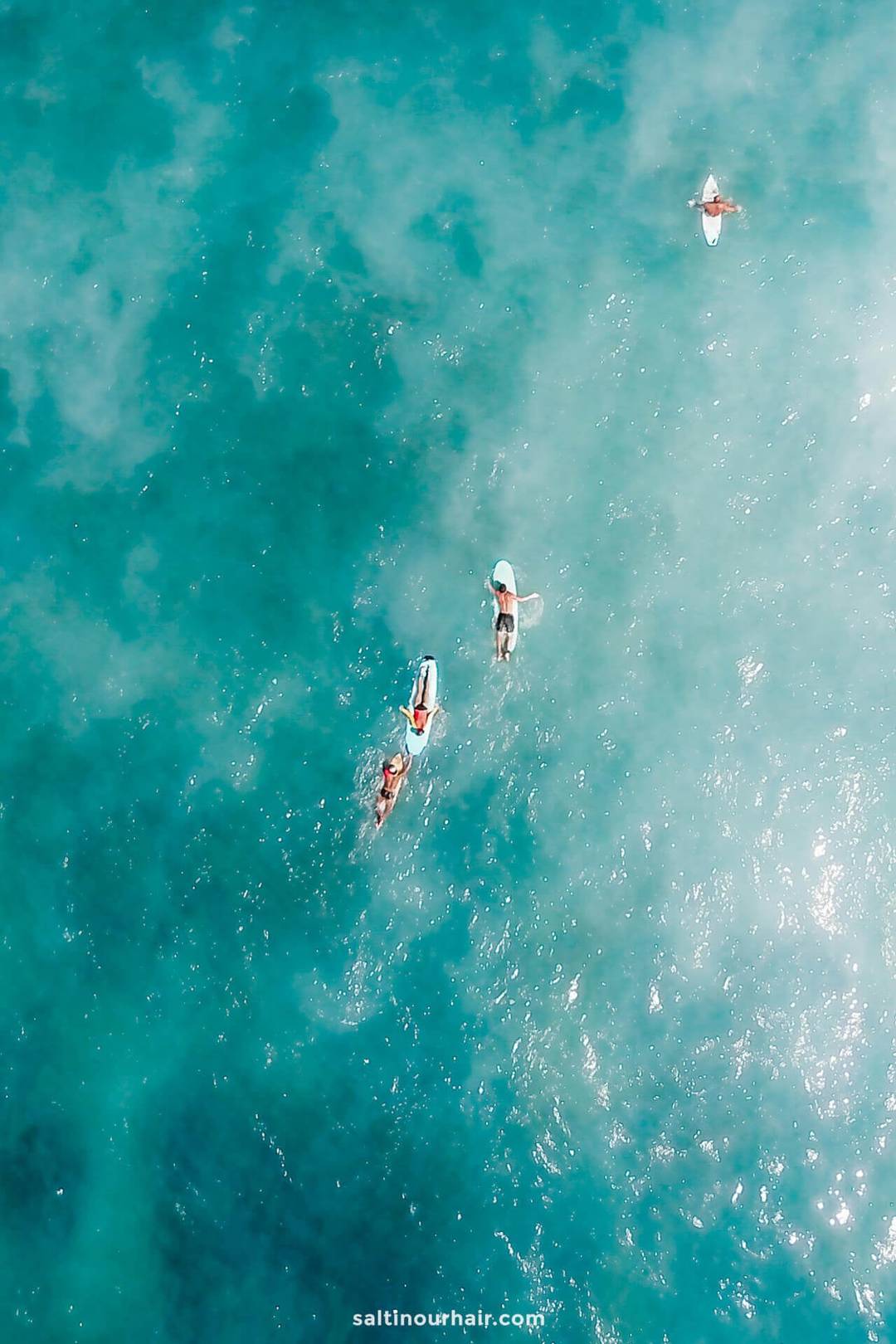 Aerial view of four surfers paddling on surfboards in the clear blue ocean water off Canggu, Bali.