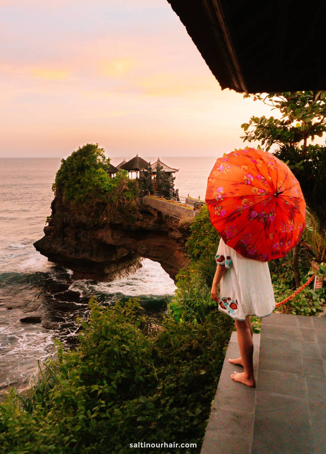 A girl holding a red floral umbrella stands on a terrace overlooking Tanah Lot Temple near Canggu, Bali, as the sun sets over the rocky outcrop.