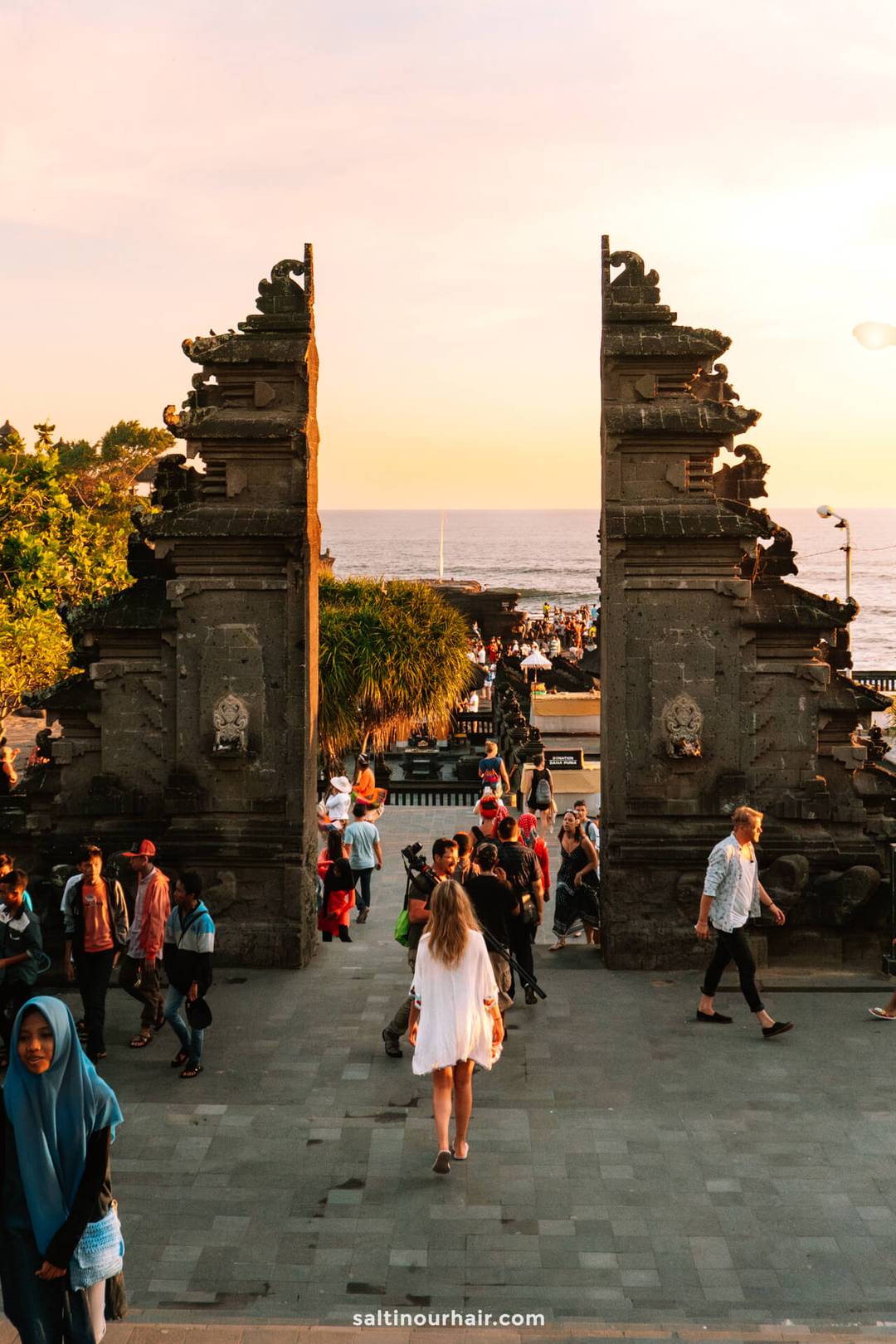 Crowds of people walk through a traditional stone gateway toward the ocean at sunset, with trees and greenery on the left side.