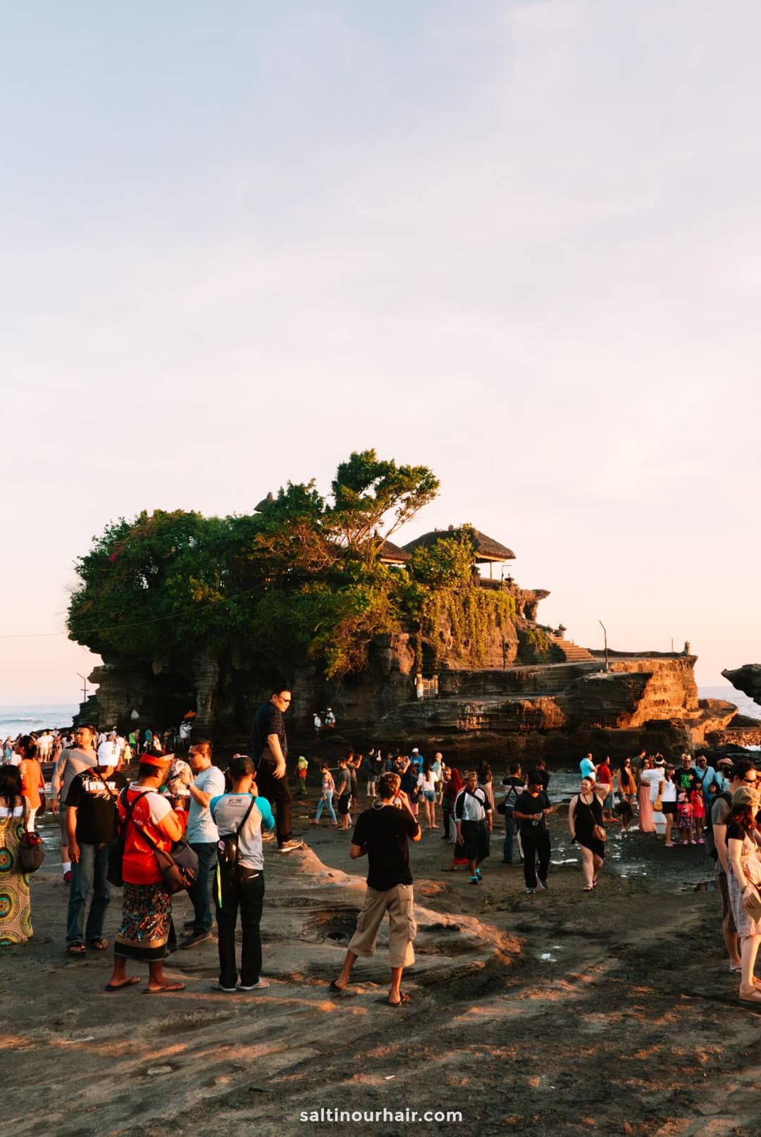 A large crowd gathers near Tanah Lot temple in Canggu, Bali, which sits atop a rocky outcrop surrounded by greenery under a partly cloudy sky.
