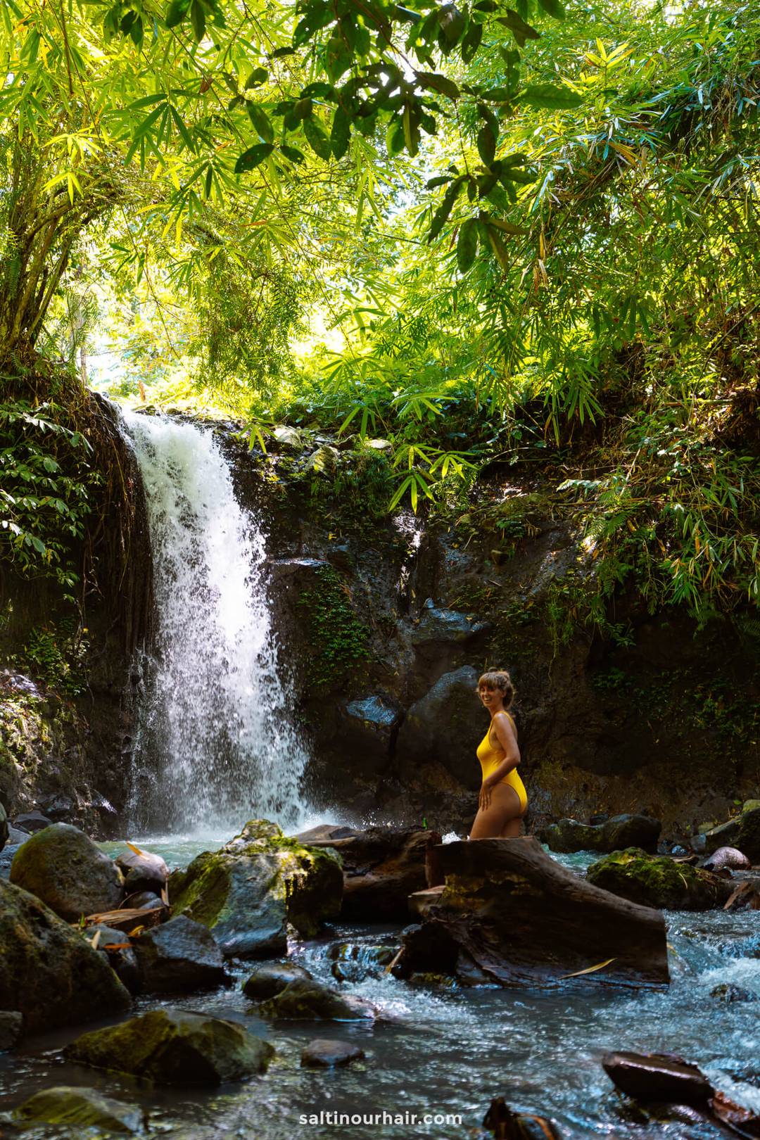 A person in a yellow swimsuit stands on rocks beside a small waterfall, surrounded by dense green foliage, capturing the tranquil beauty found near many homestay Bali retreats.