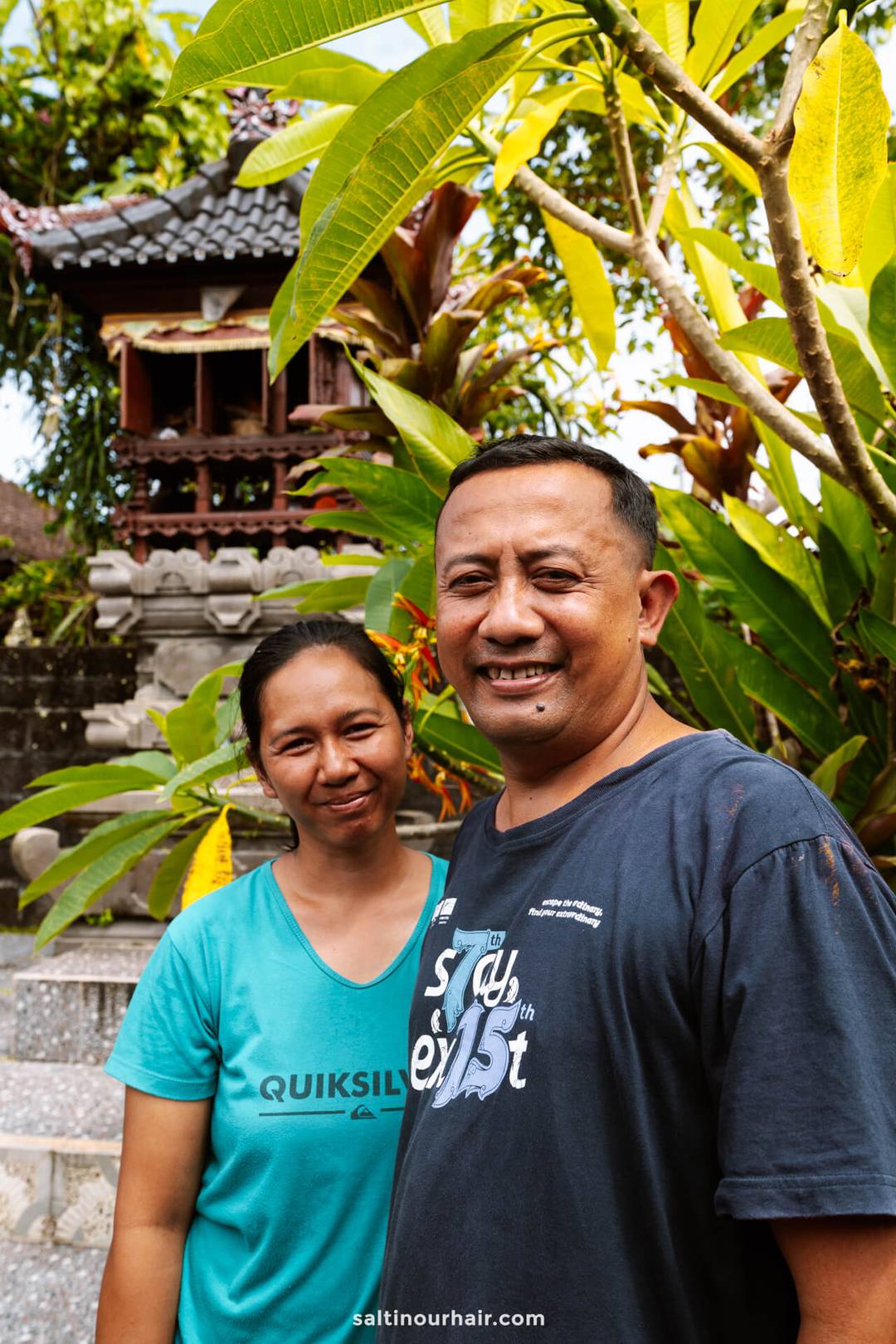 Two adults stand outdoors in front of a small traditional Balinese shrine and lush green plants, both smiling at the camera, capturing the welcoming atmosphere of a homestay Bali experience.