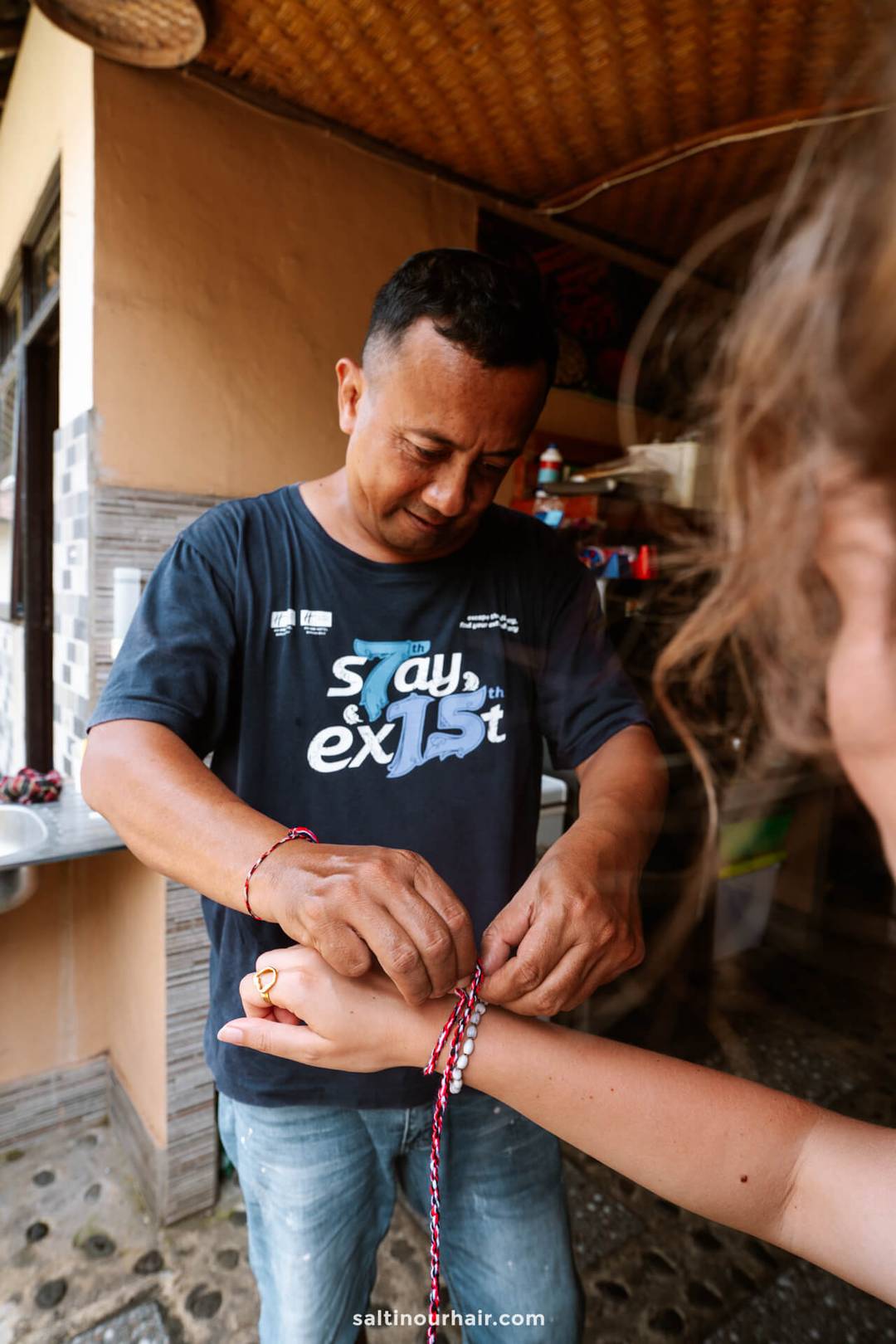A man ties a red and white string bracelet around a person&rsquo;s wrist inside a rustic homestay Bali setting.