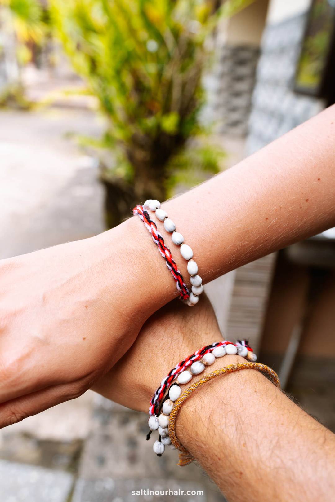 Two wrists with handmade bead and string bracelets, one wrist above the other, shown outdoors with a blurred background&mdash;capturing a relaxed homestay Bali vibe.