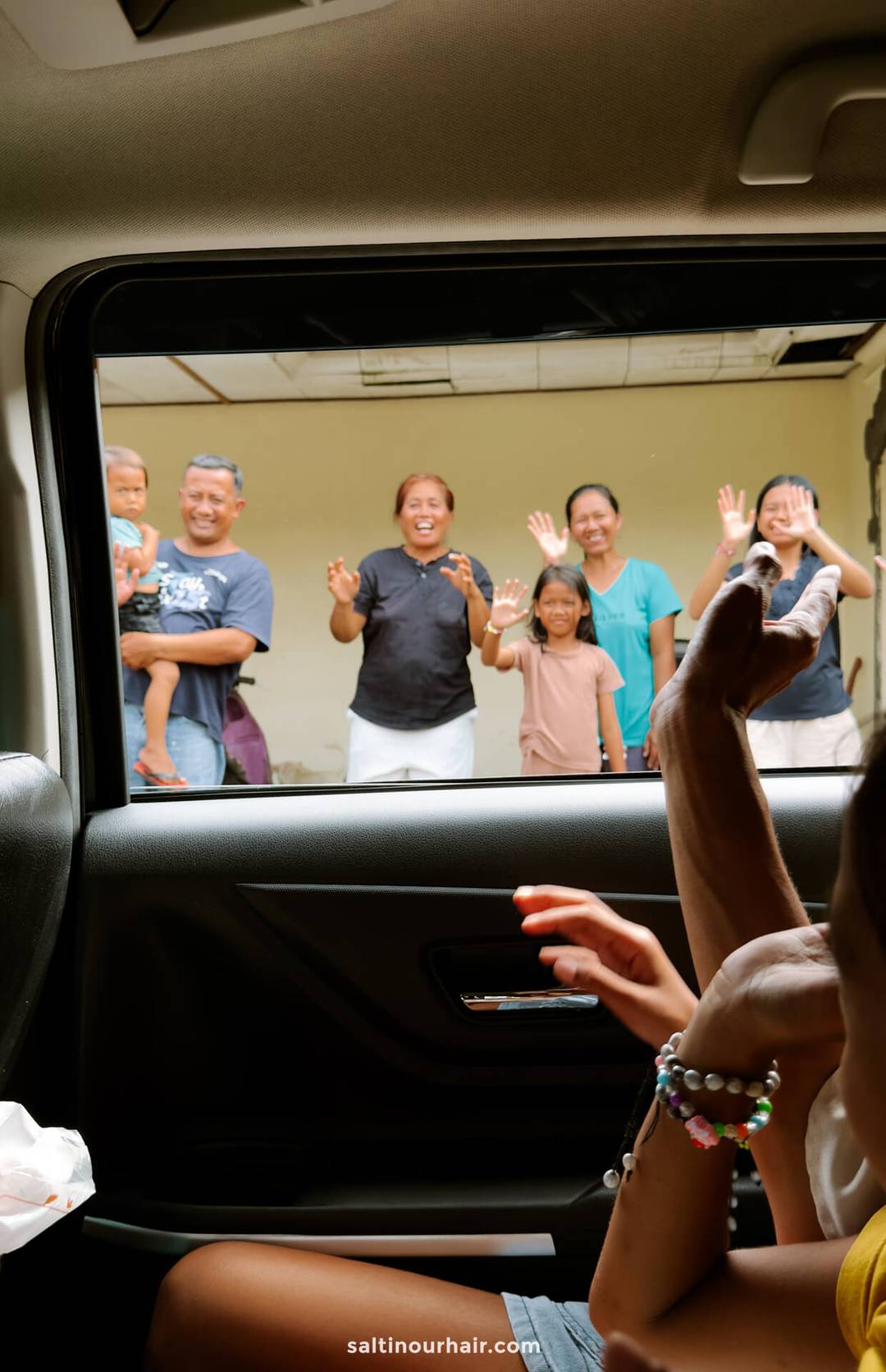 A group of six people, including children and adults, wave and smile outside a car window at their homestay Bali, while two people sit inside the car.