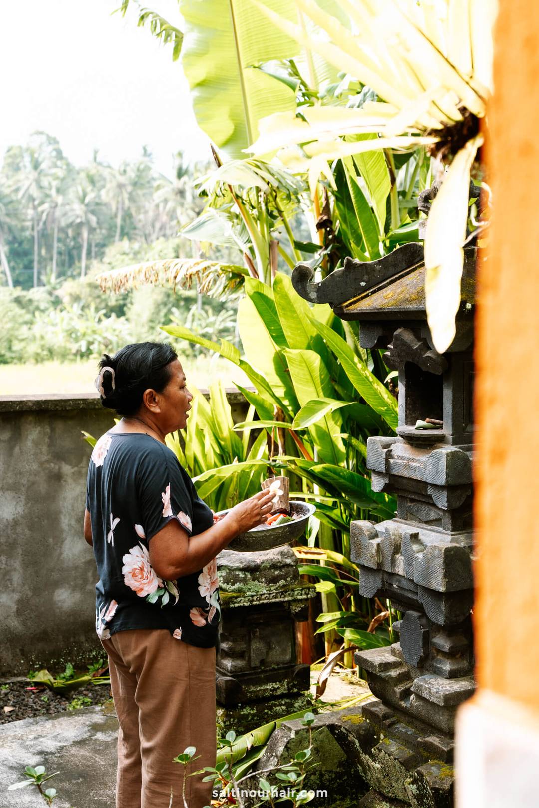 A woman stands outdoors at a homestay Bali, holding a plate and making an offering at a stone shrine surrounded by lush green plants.