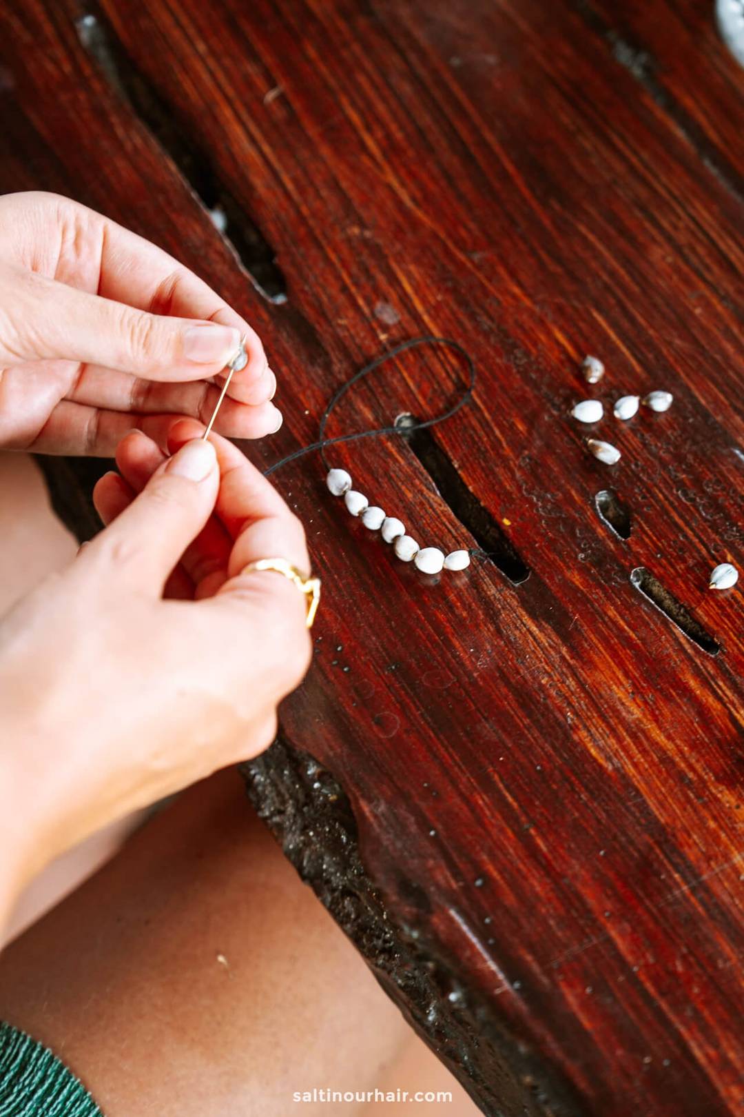 A person threads small white seashells onto a black string, crafting a shell bracelet on a wooden surface&mdash;an artisanal touch often found at a cozy homestay Bali experience.