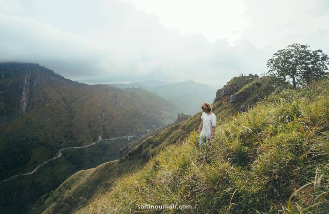 Person standing on a grassy hillside at Little Adam&rsquo;s Peak in Ella, Sri Lanka, overlooking a winding road and distant mountains under a cloudy sky.