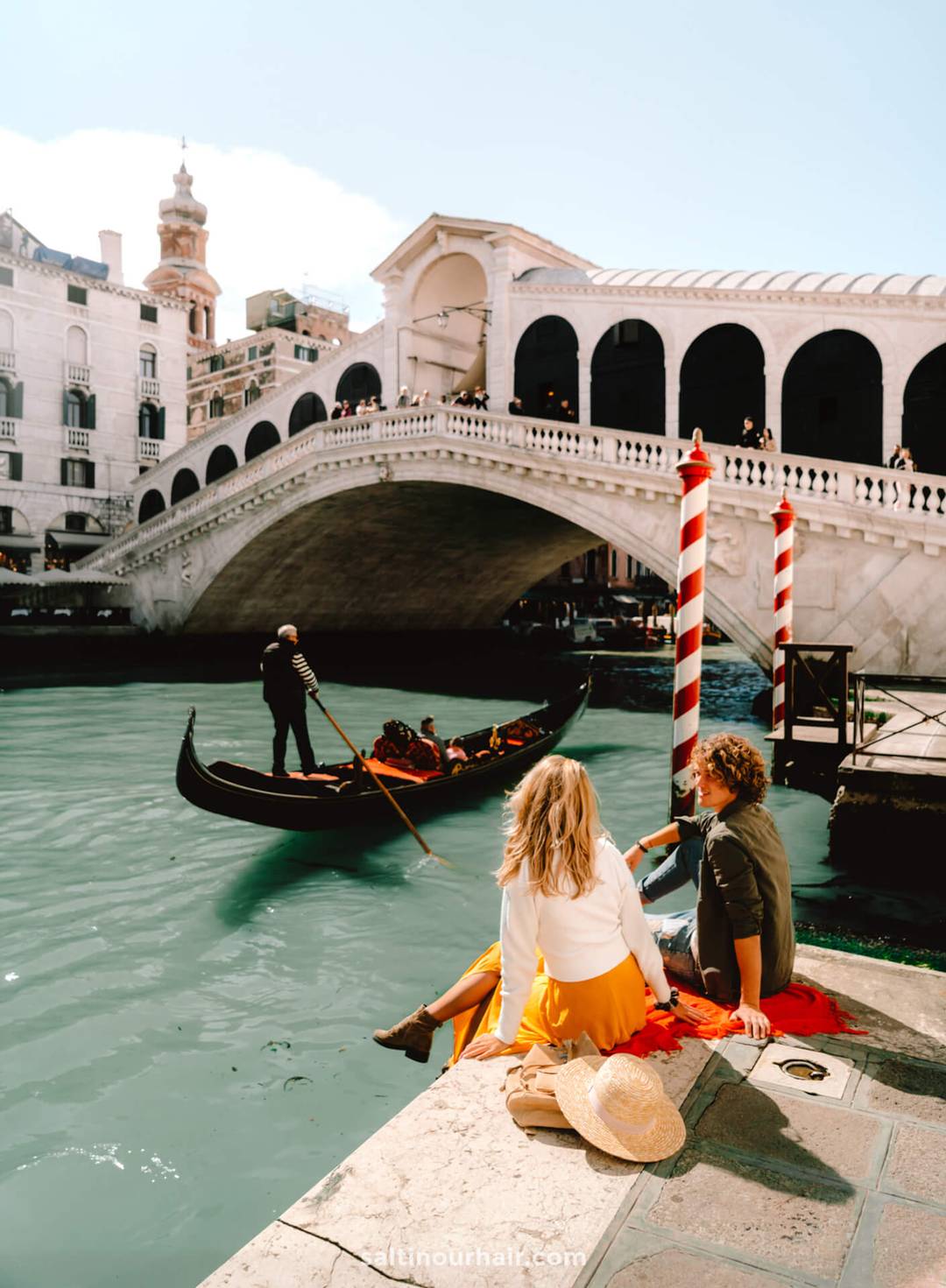 A couple sits by the canal near the Rialto Bridge in Venice, watching a gondola pass by on a sunny day&mdash;one of the world&rsquo;s most romantic honeymoon destinations.