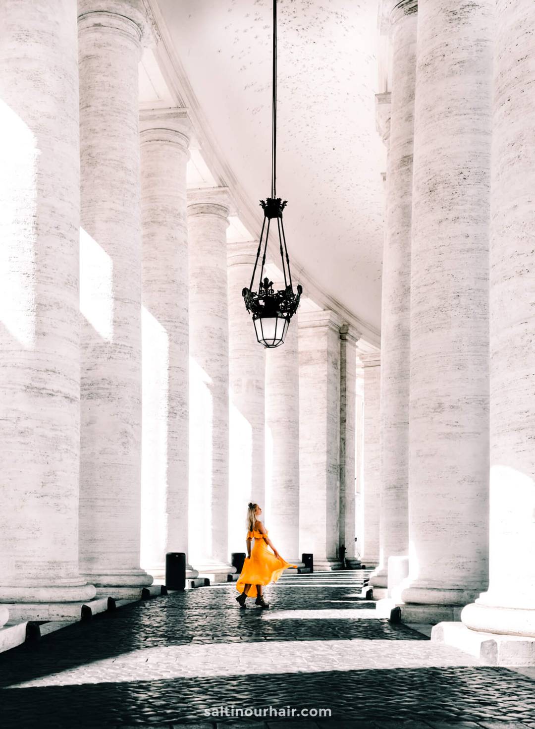 Woman in a bright yellow dress stands among tall white columns under a hanging lantern, sunlight casting strong shadows&mdash;capturing the elegance of an Italy itinerary 2 weeks journey in one striking moment.