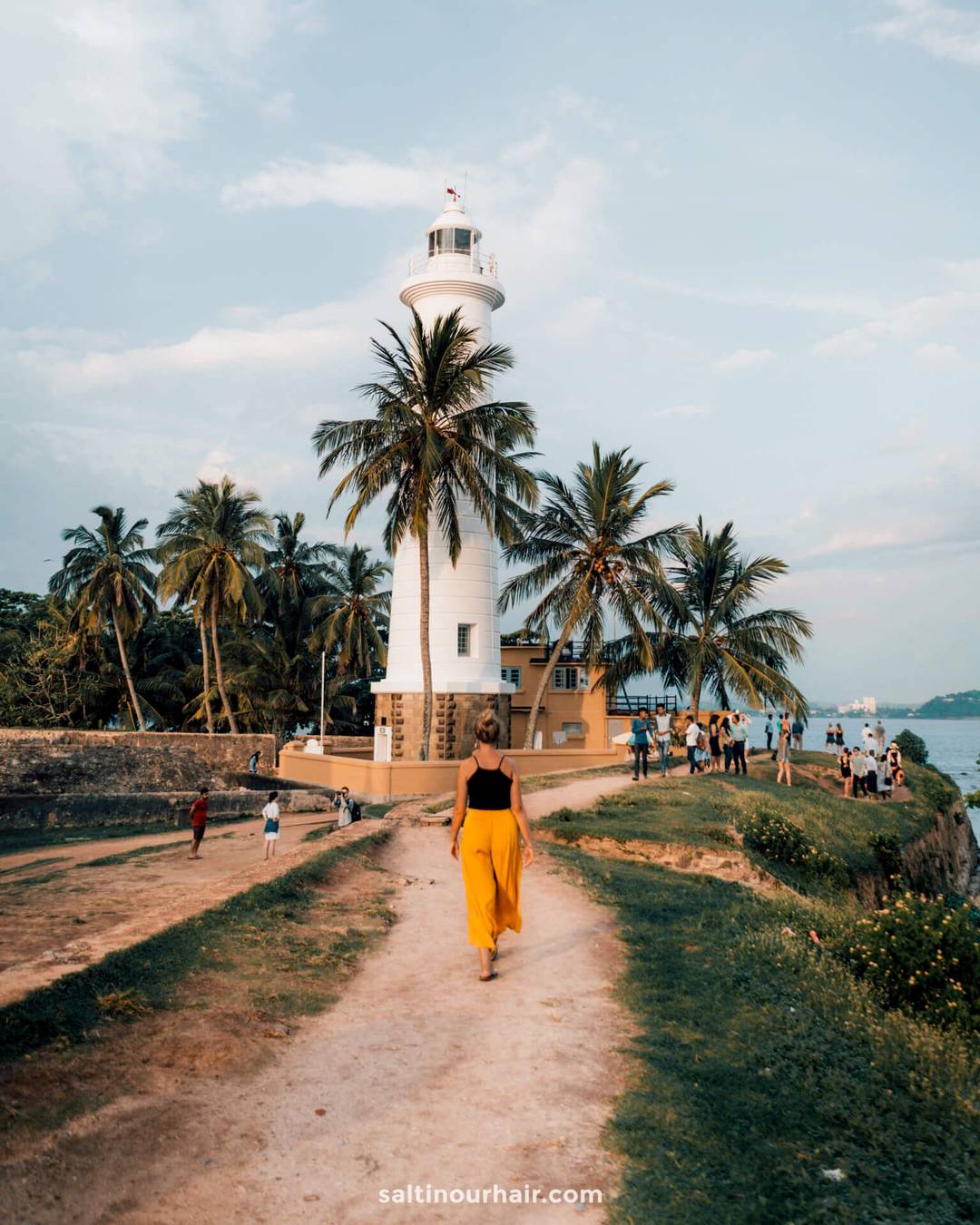Woman in yellow skirt walking toward the white Galle Lighthouse surrounded by palm trees in Sri Lanka.