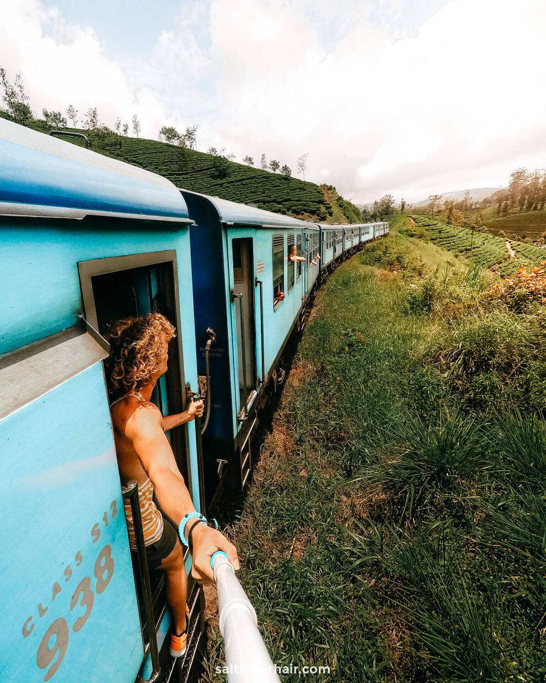 Person holding a selfie stick leans out of a blue Kandy Ella train traveling through green countryside, with open train doors and the curved track visible.