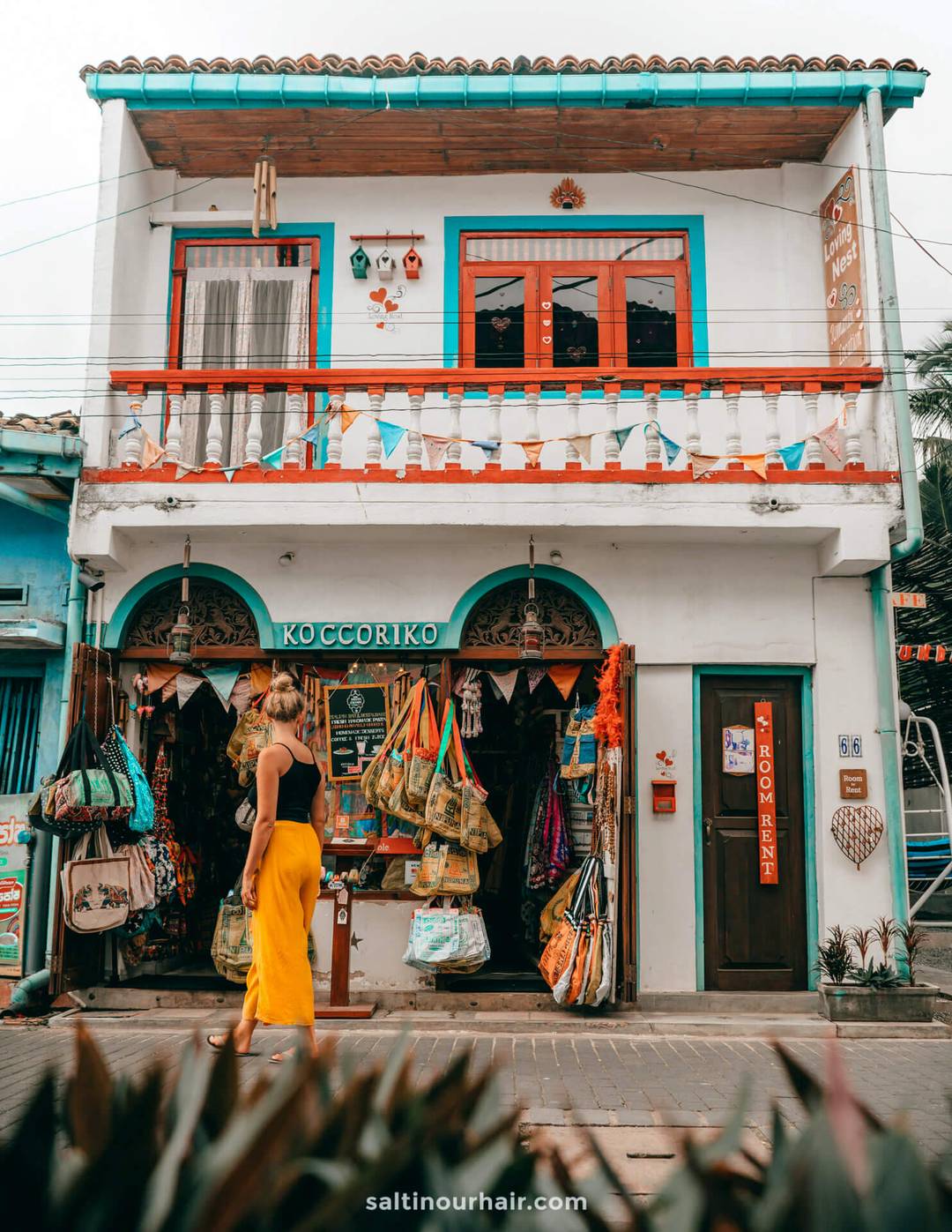 Woman in yellow skirt browsing Koccoriko souvenir shop in Galle, Sri Lanka, with colorful bunting and tote bags displayed outside.