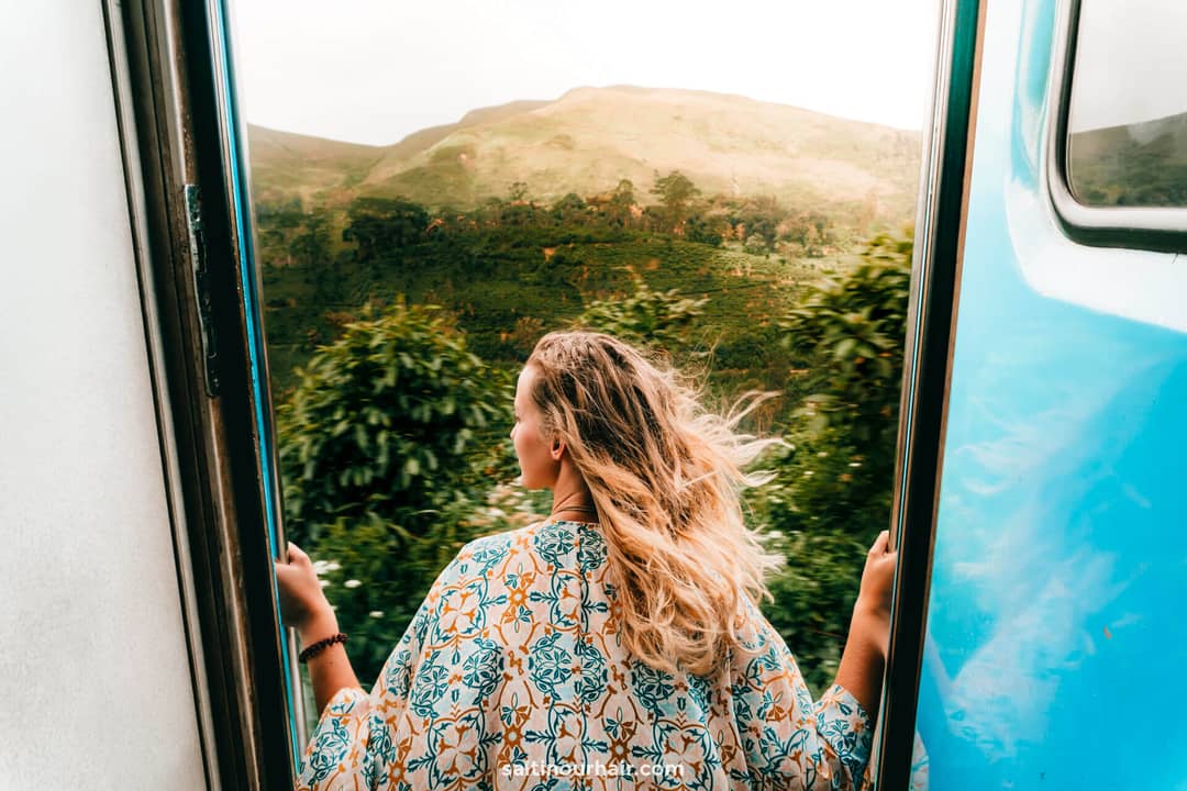 A person with long blonde hair leans out of the open doorway of a blue train during the scenic Sri Lanka Ella to Kandy train ride, admiring the lush green hills and trees in the background.
