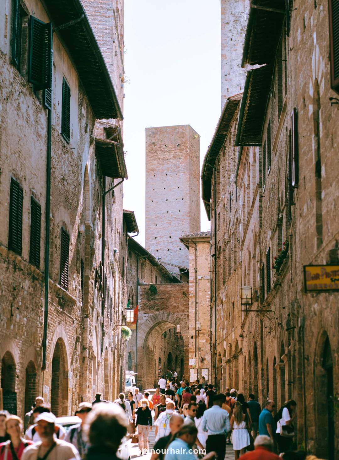A narrow stone street in the historic town of San Gimignano, Tuscany, Italy, is crowded with people, with tall medieval buildings and a tower rising in the background.