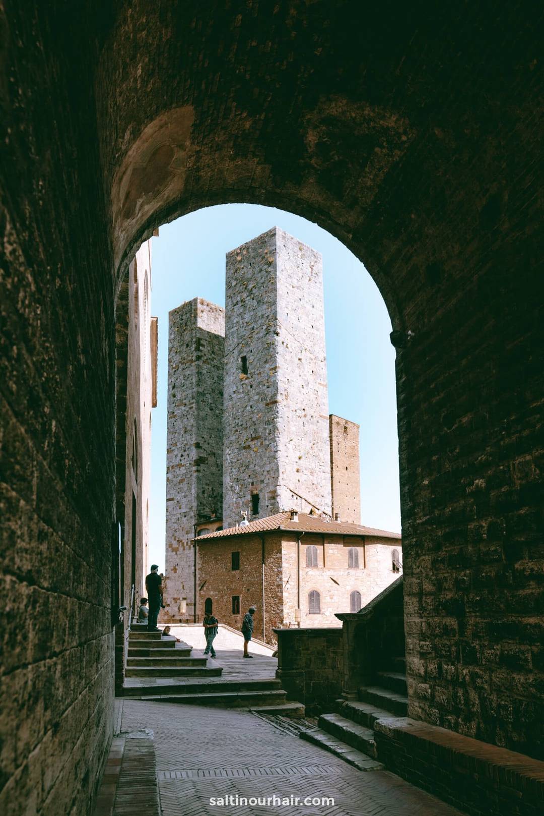 View through an arched stone doorway of tall medieval towers and a brick building in San Gimignano, Tuscany, Italy, with a few people walking on the steps.