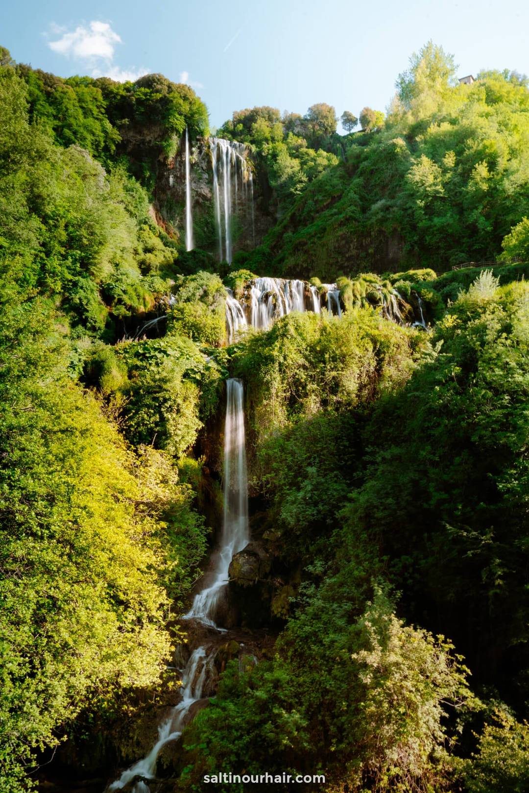 A tall, multi-tiered waterfall flows down lush green cliffs surrounded by dense foliage under a clear blue sky&mdash;one of the breathtaking things to do on a 2-week italy itinerary