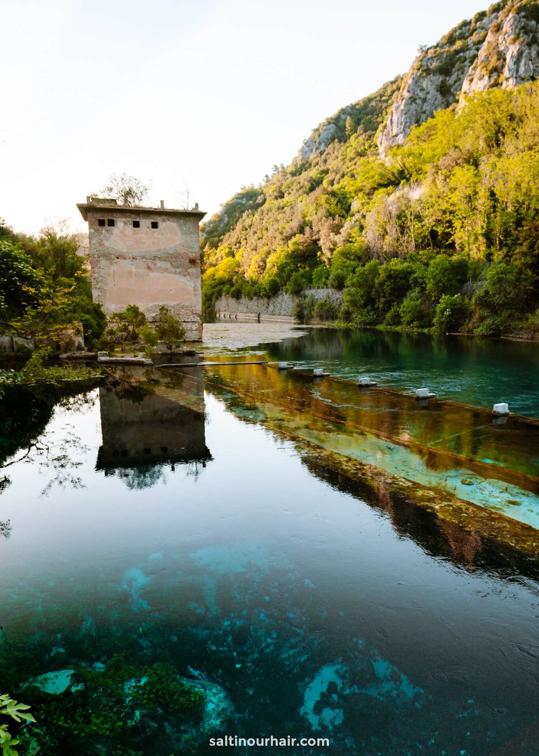 A rectangular stone building stands by a clear blue river in Umbria, Italy, surrounded by lush green trees and rocky hills&mdash;a tranquil scene perfect for your list of things to do in Umbria Italy&mdash;its reflection shimmering in the water.