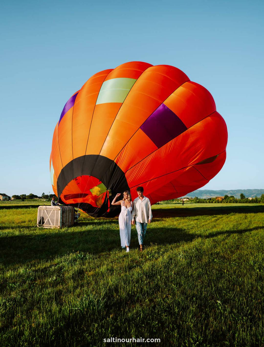 A couple walks on grass near a partially deflated hot air balloon with a bright red and orange envelope&mdash;a picturesque moment in a field under a clear blue sky, showcasing unique things to do in Umbria, Italy.