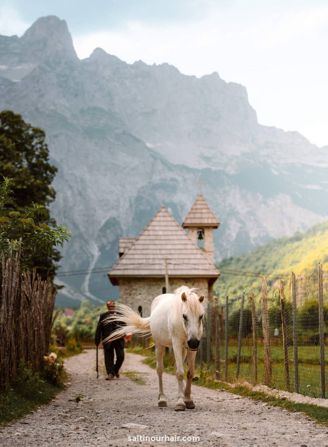 A white horse walks along a dirt path near a small stone building in Theth, Albania, with mountains rising behind and a person following—an idyllic scene perfect for your 7 day albania itinerary.
