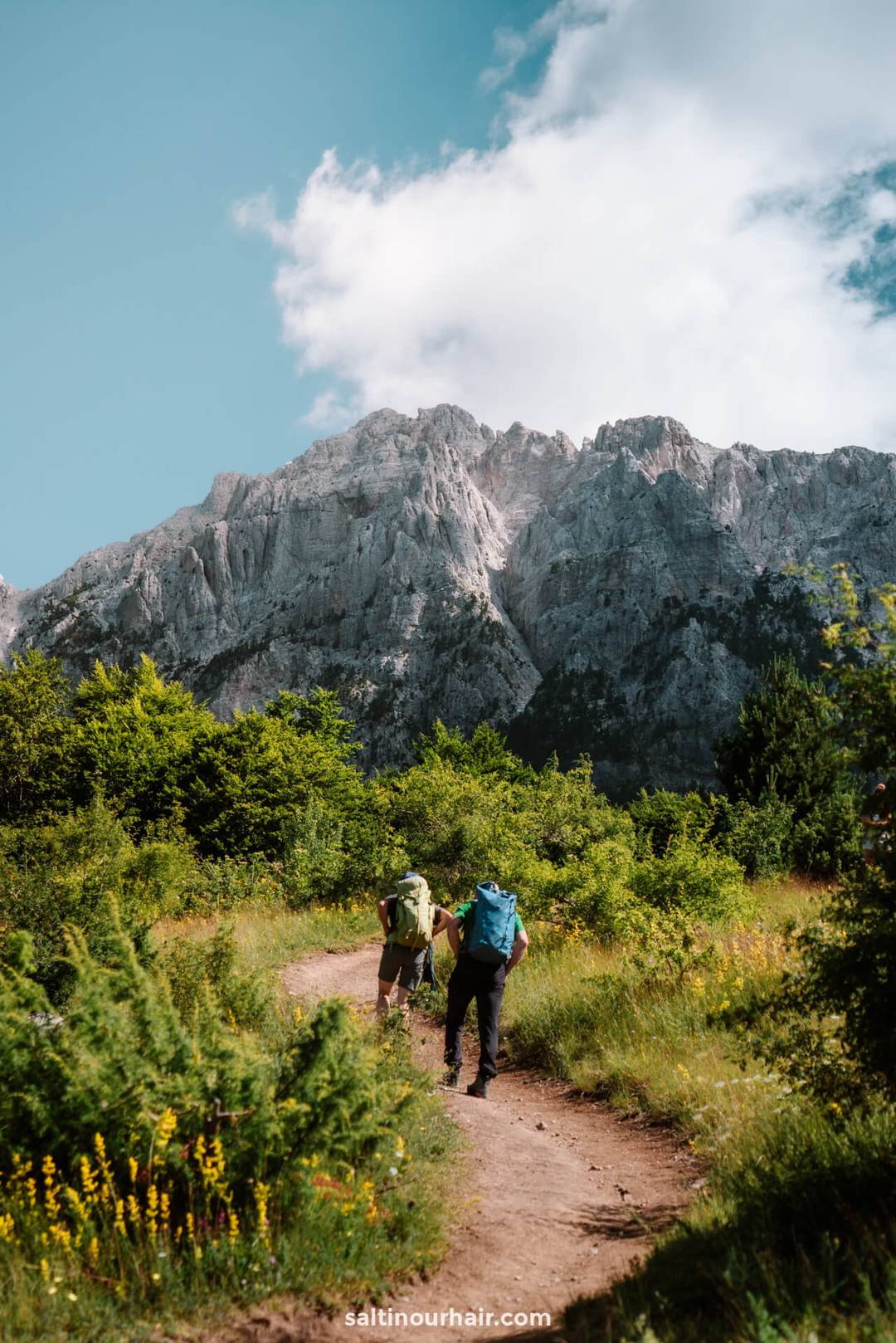 Two hikers with backpacks walk along a dirt trail surrounded by greenery, heading toward the majestic Valbona Peak in Albania under a partly cloudy sky.