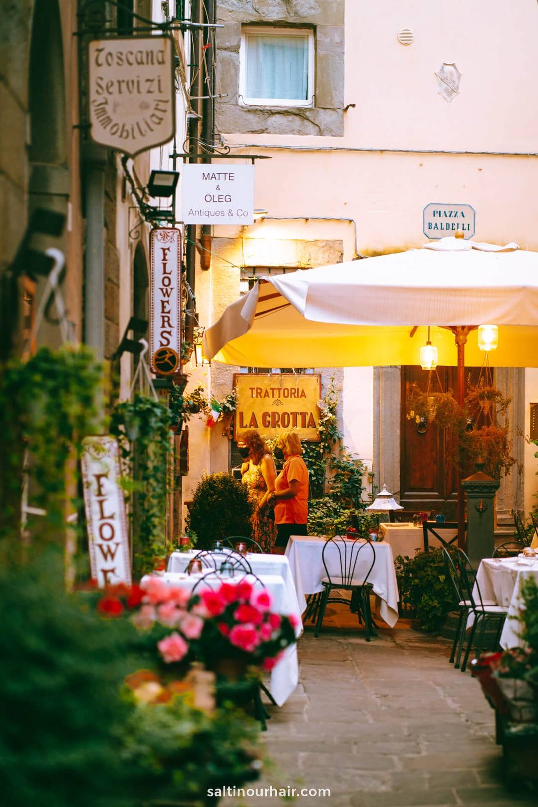 Outdoor Italian restaurant with white tablecloths, potted flowers, and warm lighting; two people speak under a large umbrella near a sign reading Trattoria La Grotta in Cortona, Tuscany, Italy.