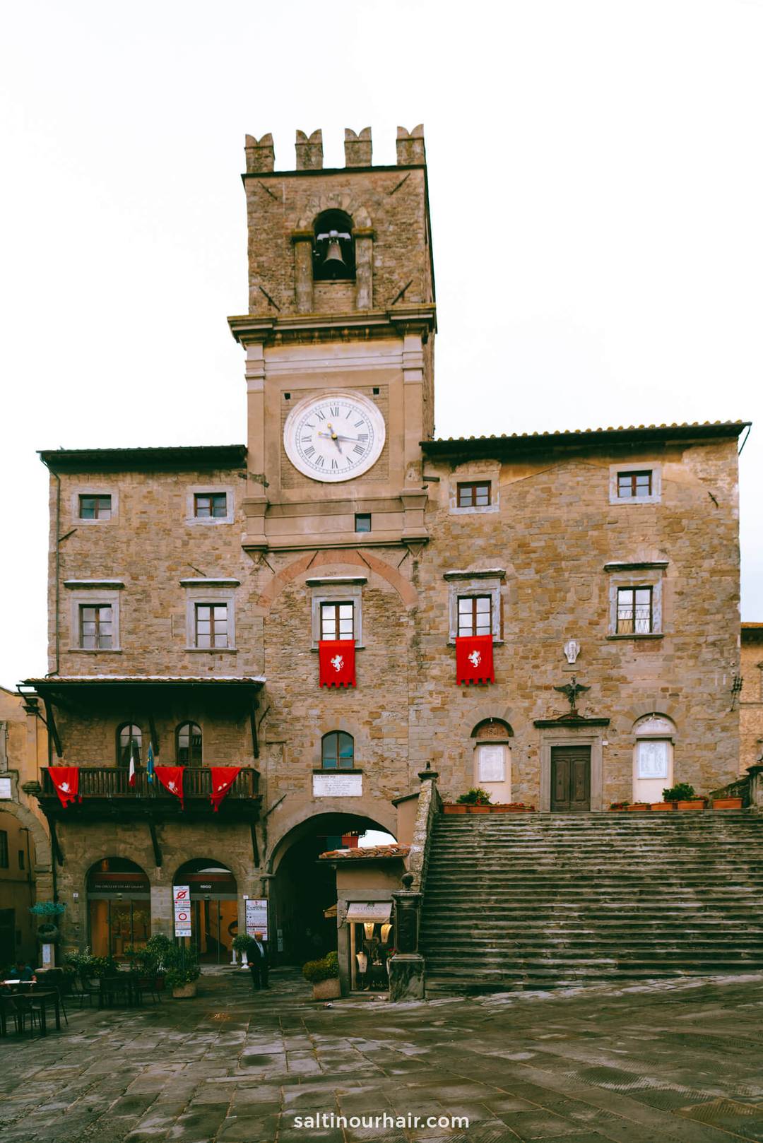 Stone clock tower building with arched entrance, large clock face, red banners, and outdoor stairway in an old European town square, typical of Tuscany, Italy.